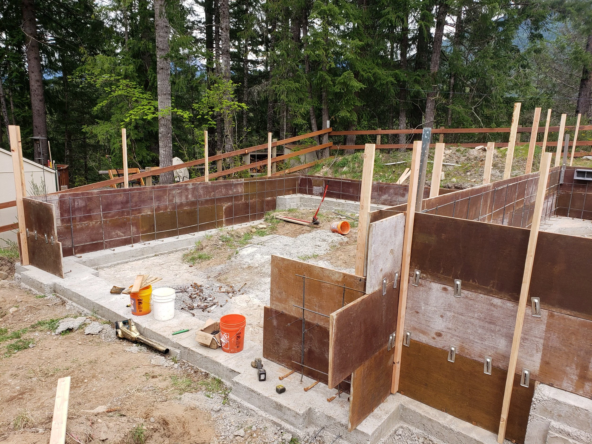 Construction site of a foundation with wooden forms and rebar, surrounded by trees in a wooded area.