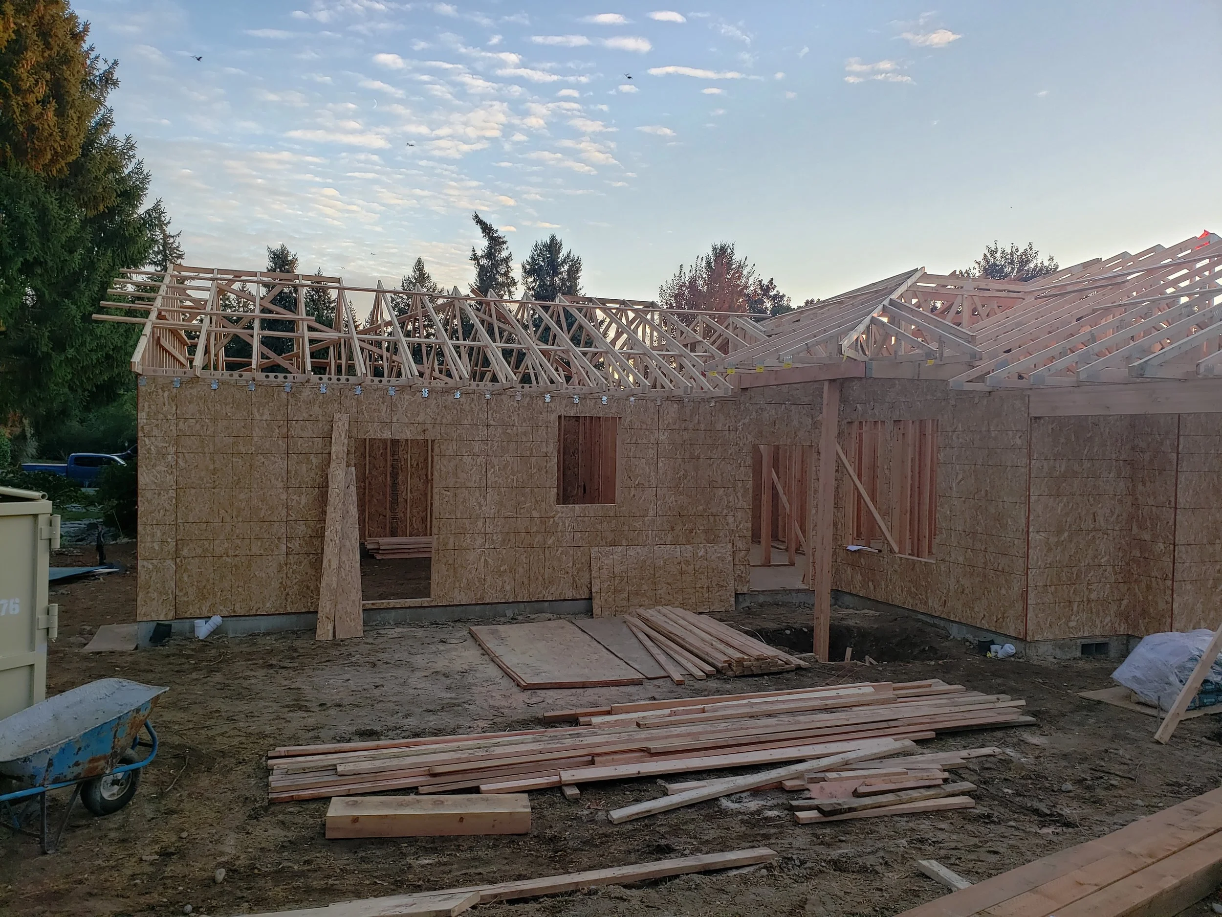 Under-construction house with exposed wooden framework and roof trusses, plywood walls, and construction materials scattered on the ground.