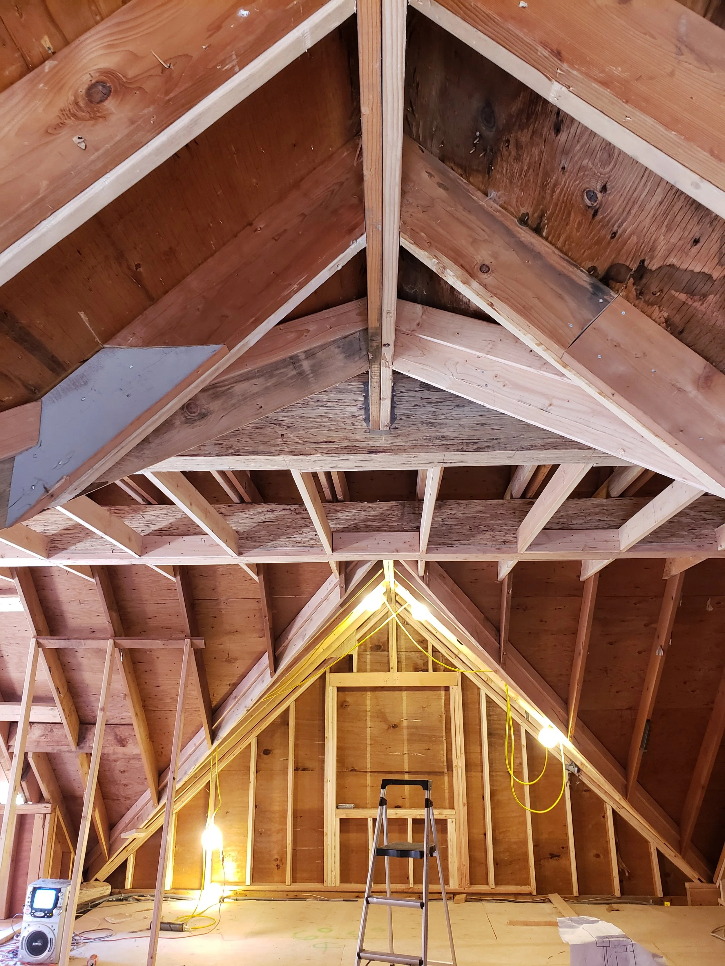 Attic construction with exposed wooden framing and insulation, with work lights and a step ladder.