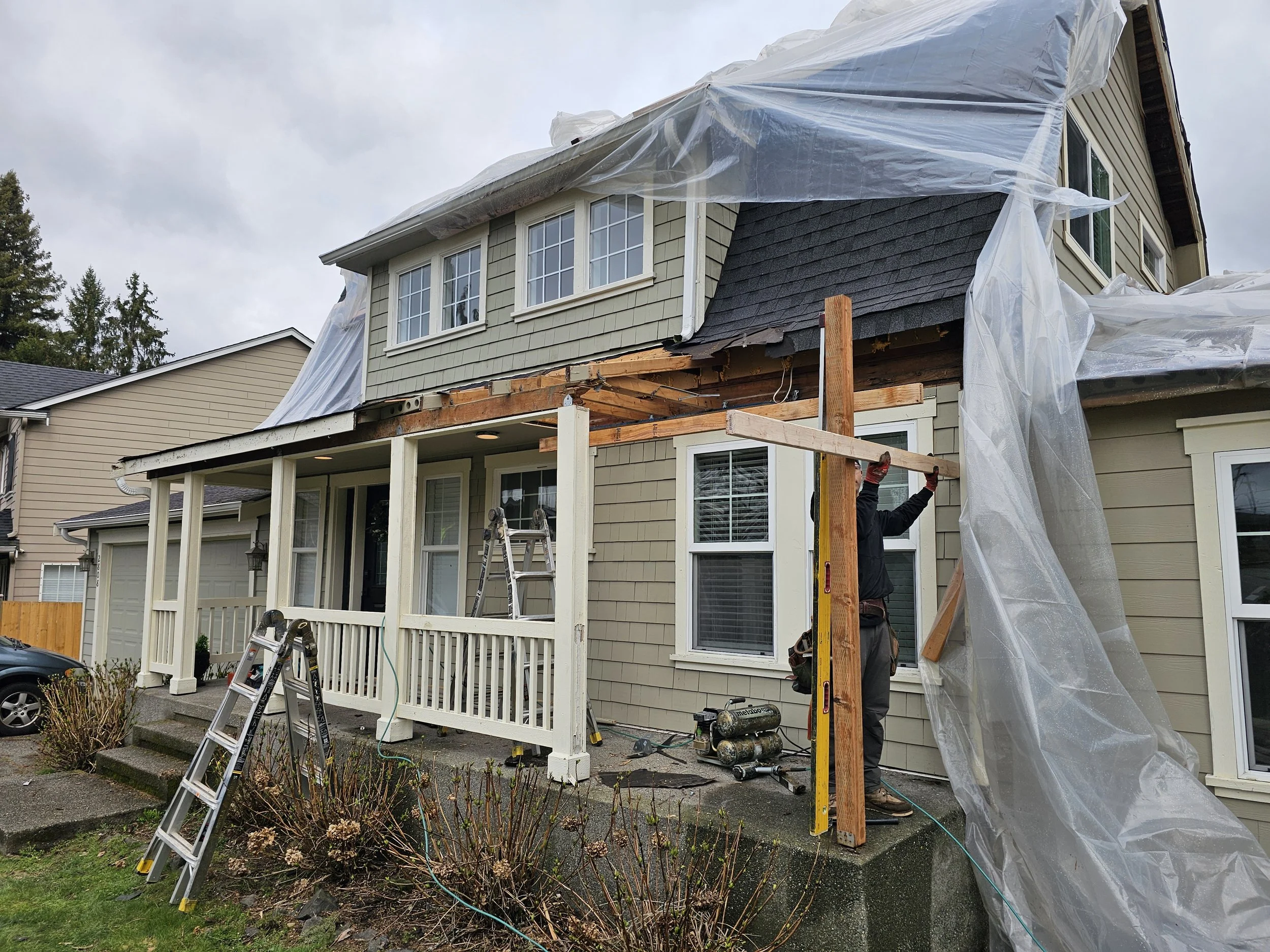Construction workers are building or renovating the porch of a house, with plastic sheeting covering parts of the house and ladders nearby.