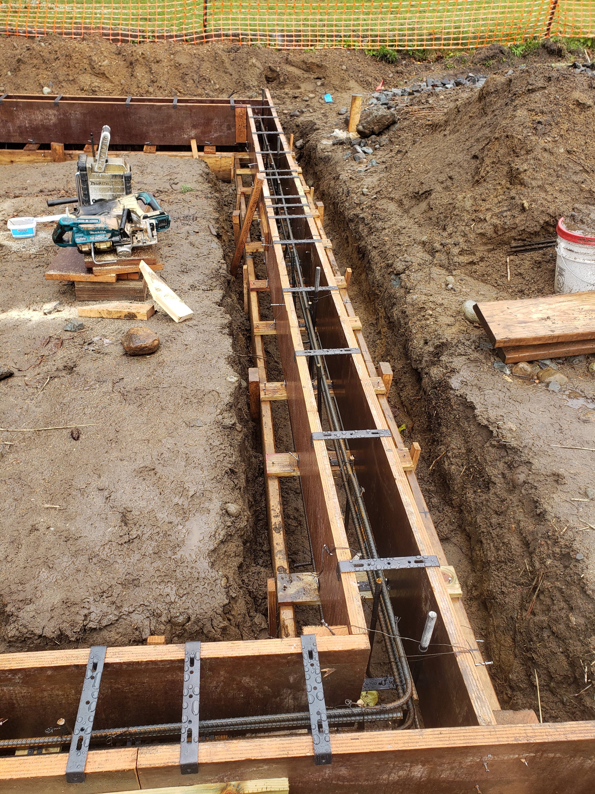 Construction site with wooden formwork and steel reinforcement for a foundation or footing, with construction tools and materials nearby.