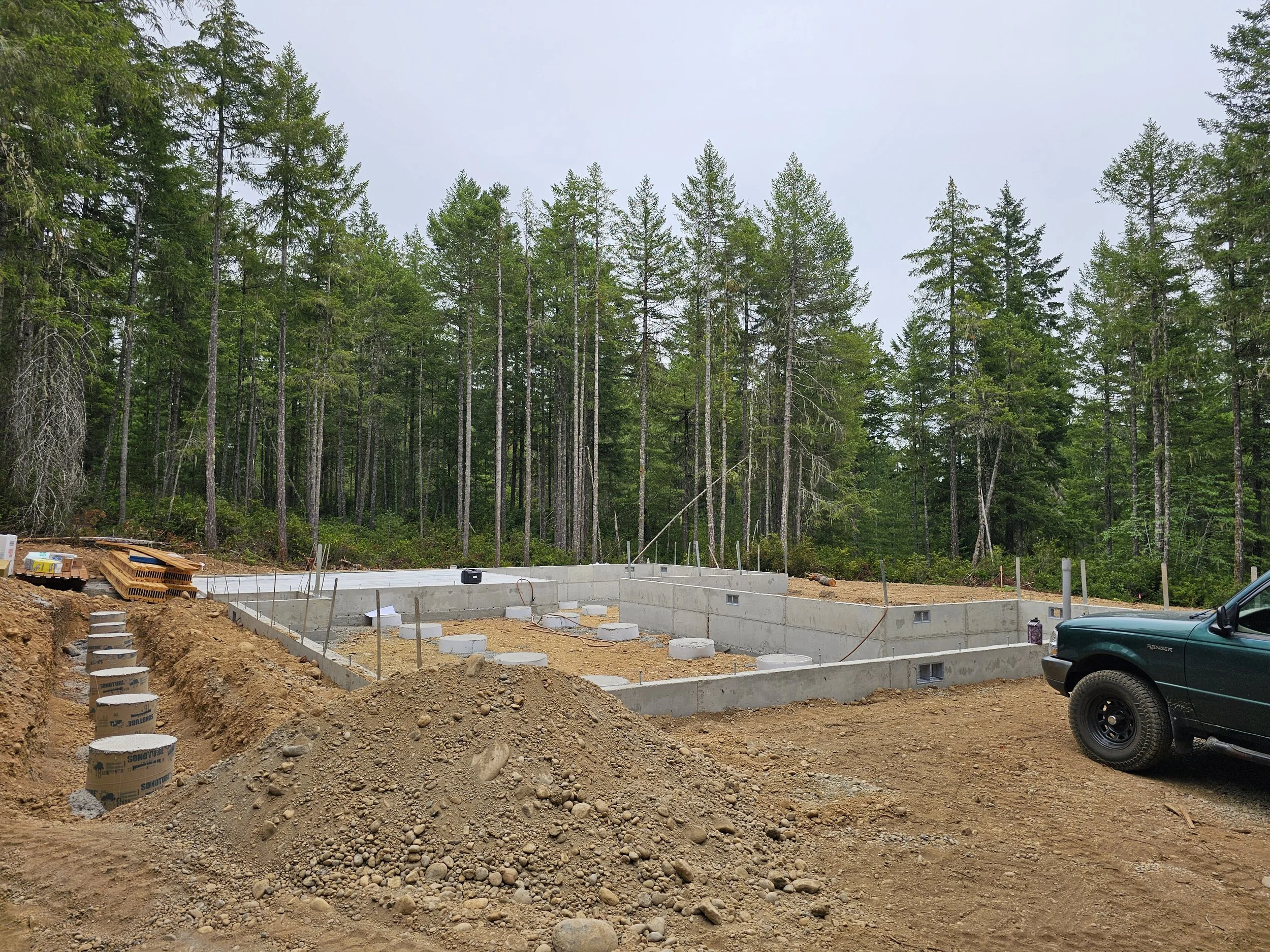 Construction site with concrete foundation, dirt piles, and a green pickup truck, surrounded by tall pine trees under an overcast sky.