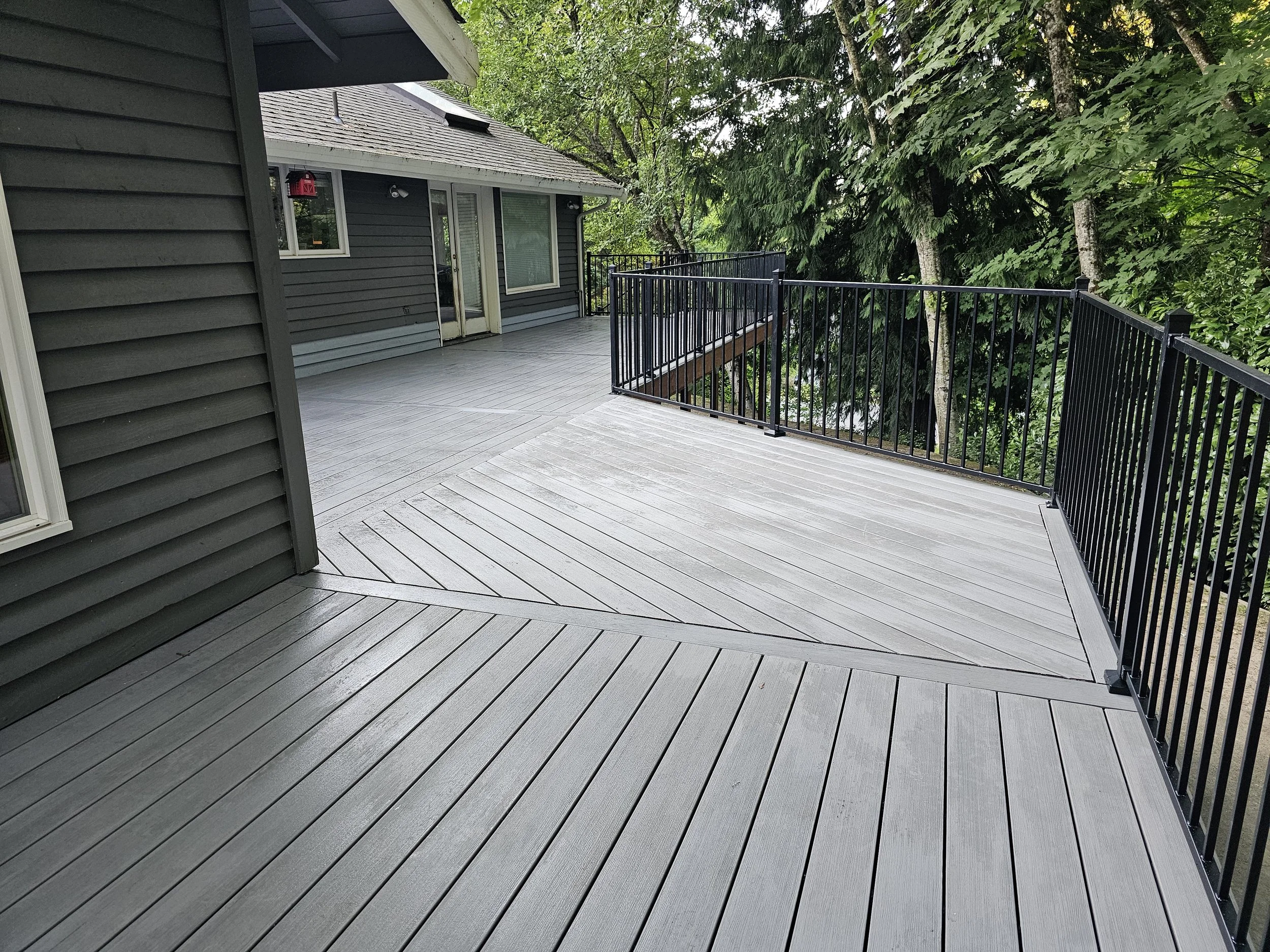 An outdoor deck with black metal railing, attached to a house with dark gray siding, surrounded by lush green trees.