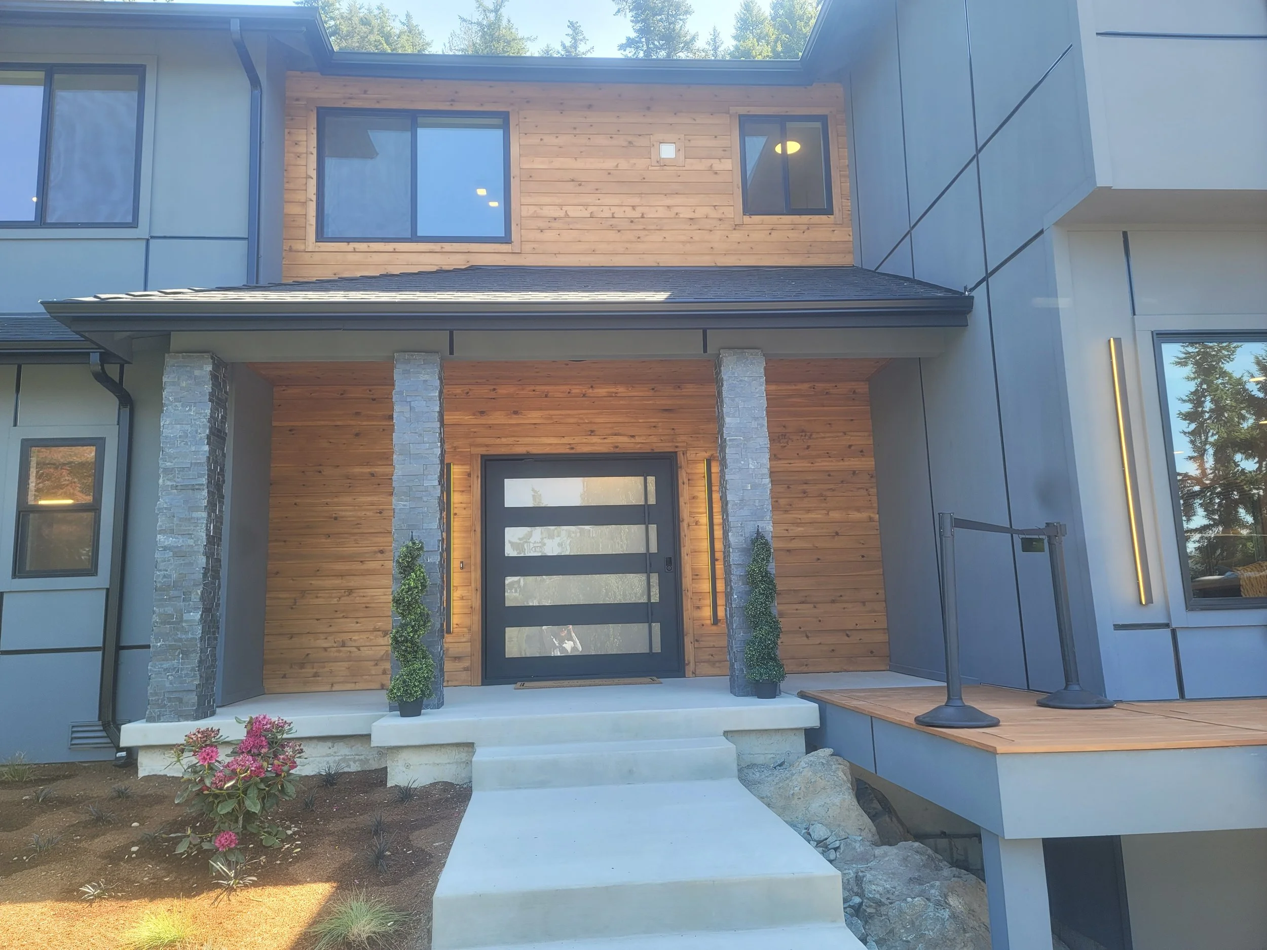 Modern custom house build with wooden and gray exterior, stone pillars, a black front door, and potted plants on the porch, with steps leading up to the entrance.