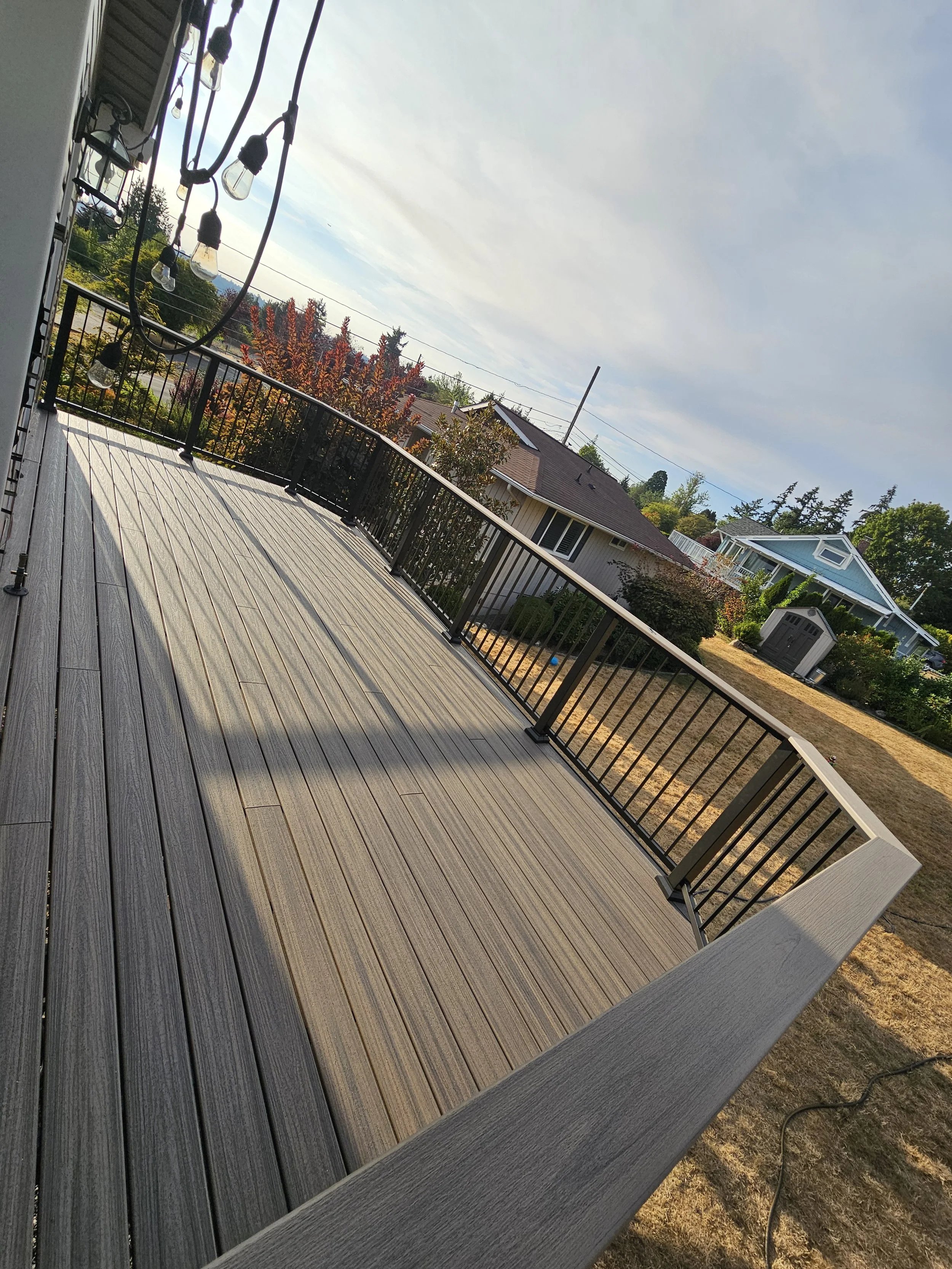A new deck build with black metal railing, outdoor string lights hanging on the side, in a residential neighborhood with houses, trees, and a cloudy sky.