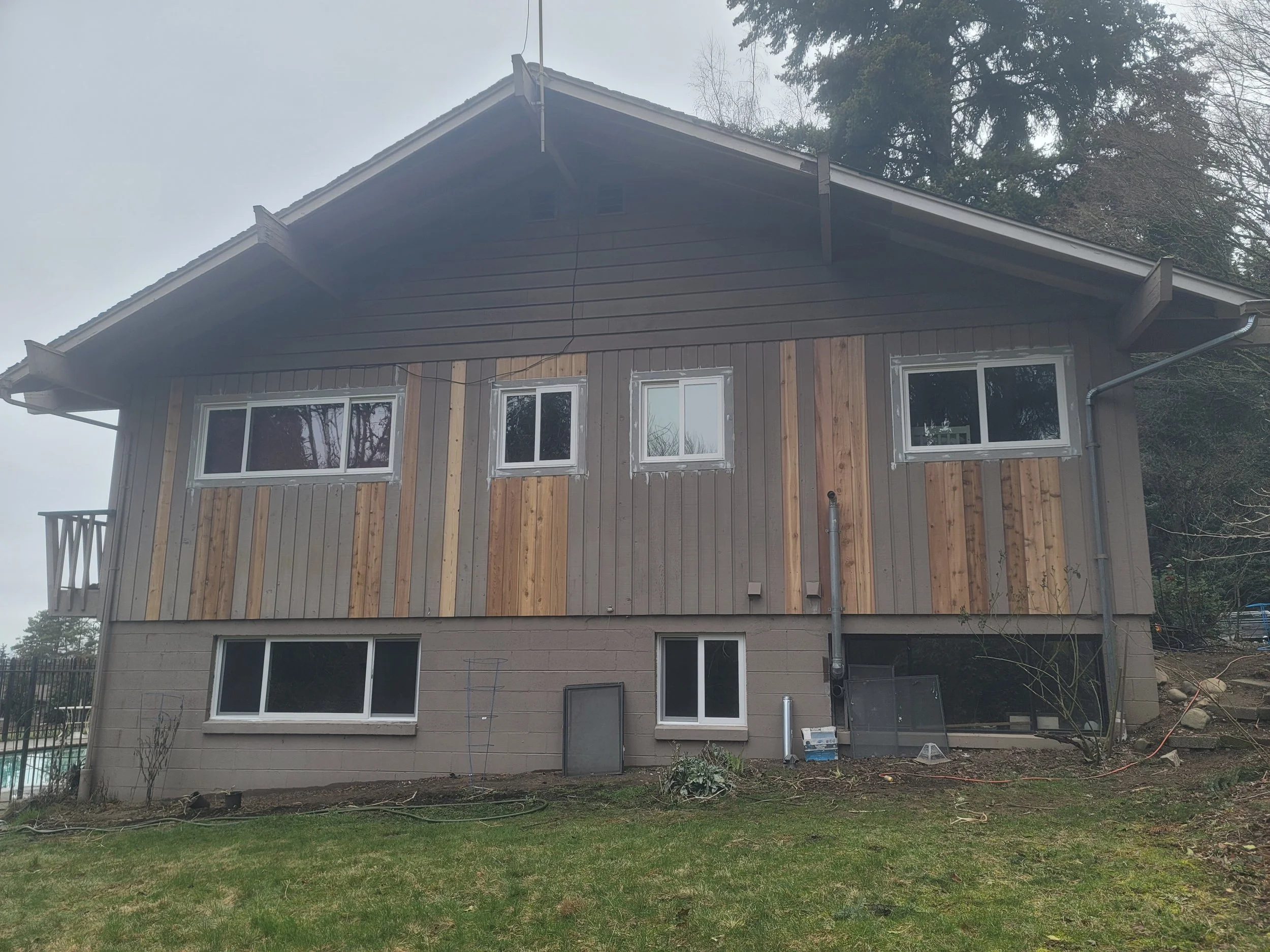 The rear of a house under renovation with a partially completed wooden siding, multiple windows, a small balcony, and stairs on the right side leading up the yard.