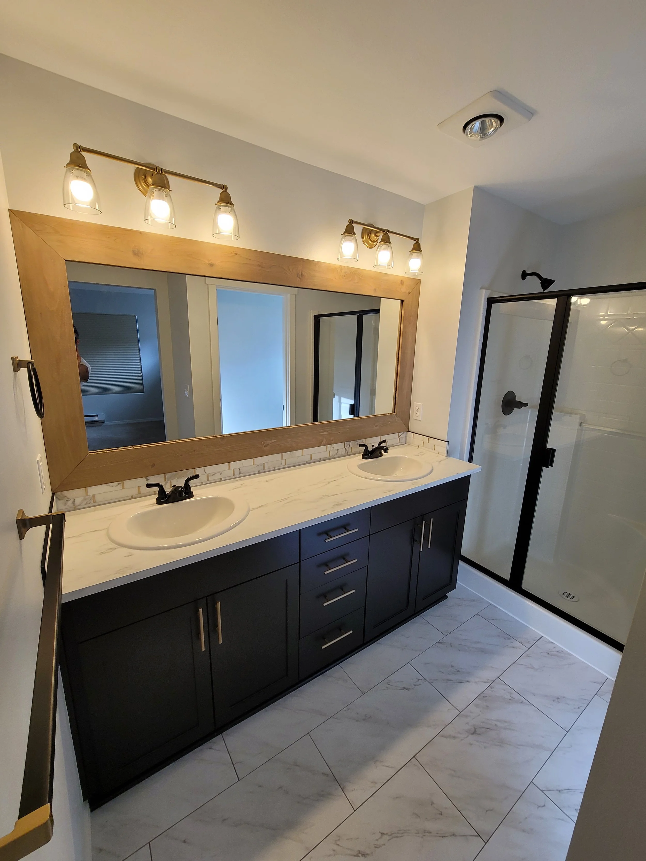A master bathroom remodel with double vanity  black cabinets, large mirror, and dual sinks; shower stall with glass door.