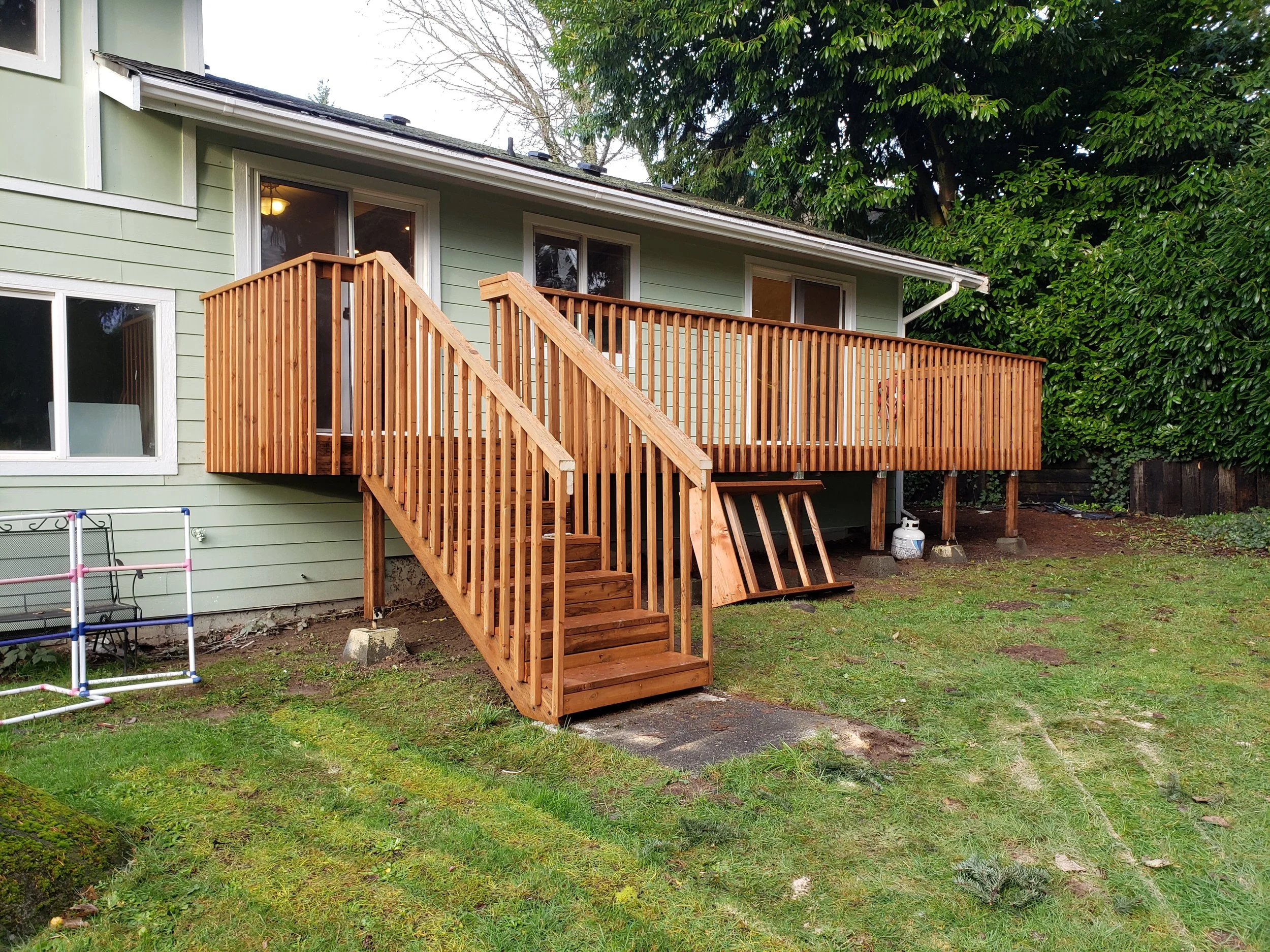 Back of a light green house with a new wooden deck and stairs, set in a grassy yard with trees and shrubs.
