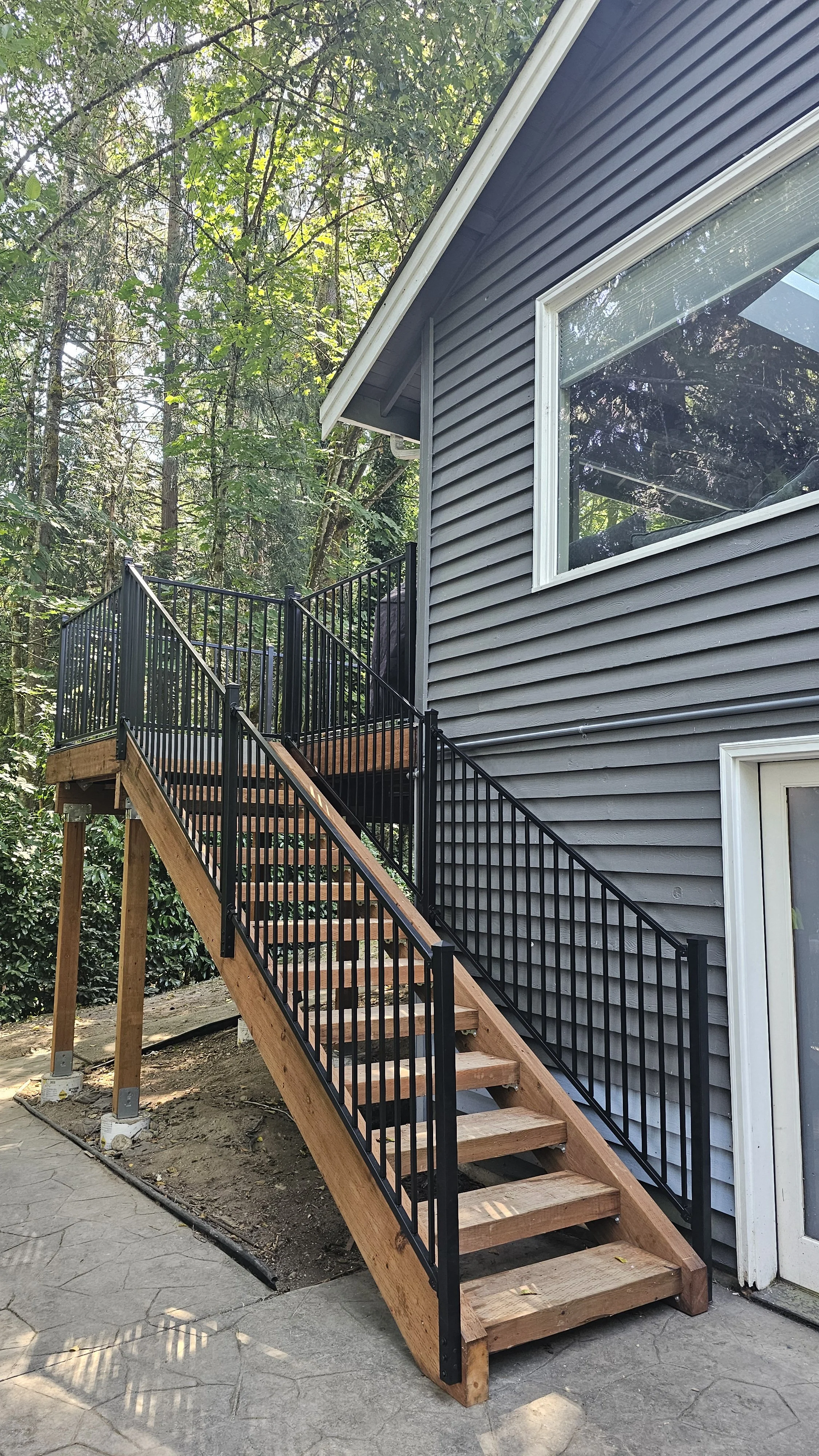 Exterior view of a house with a newly built wooden staircase with black metal railings leading to a second-floor porch surrounded by trees.