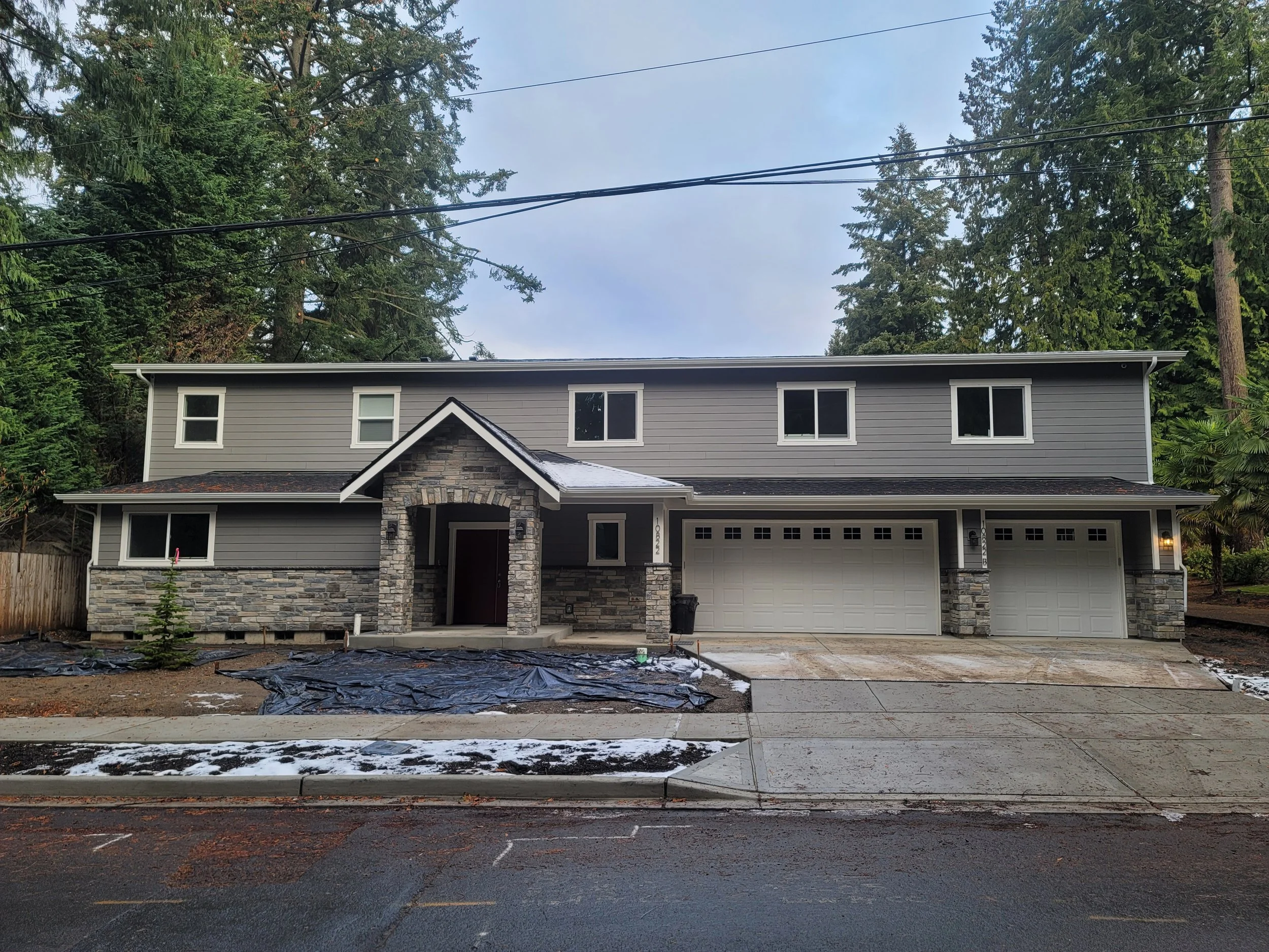 Newly constructed custom build two-story house with gray siding, stone accents, and a front porch, situated on a paved street in a wooded area.