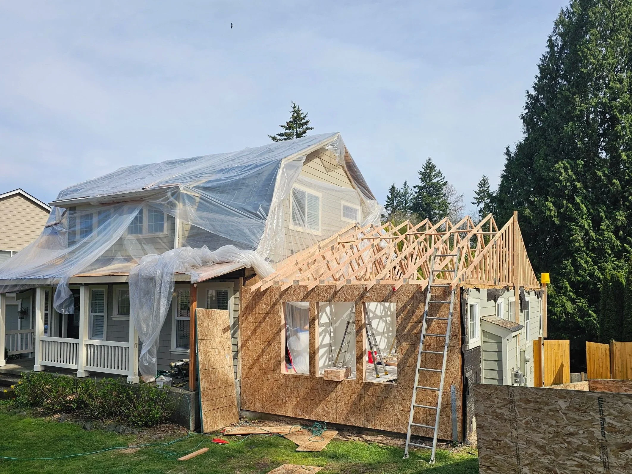 A house under construction with wooden framing and plywood sheathing, plastic sheeting covering the upper part, surrounded by trees and a partly cloudy sky.