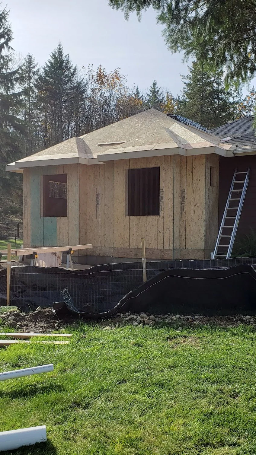 A house addition under construction, surrounded by a grassy yard and trees in the background.