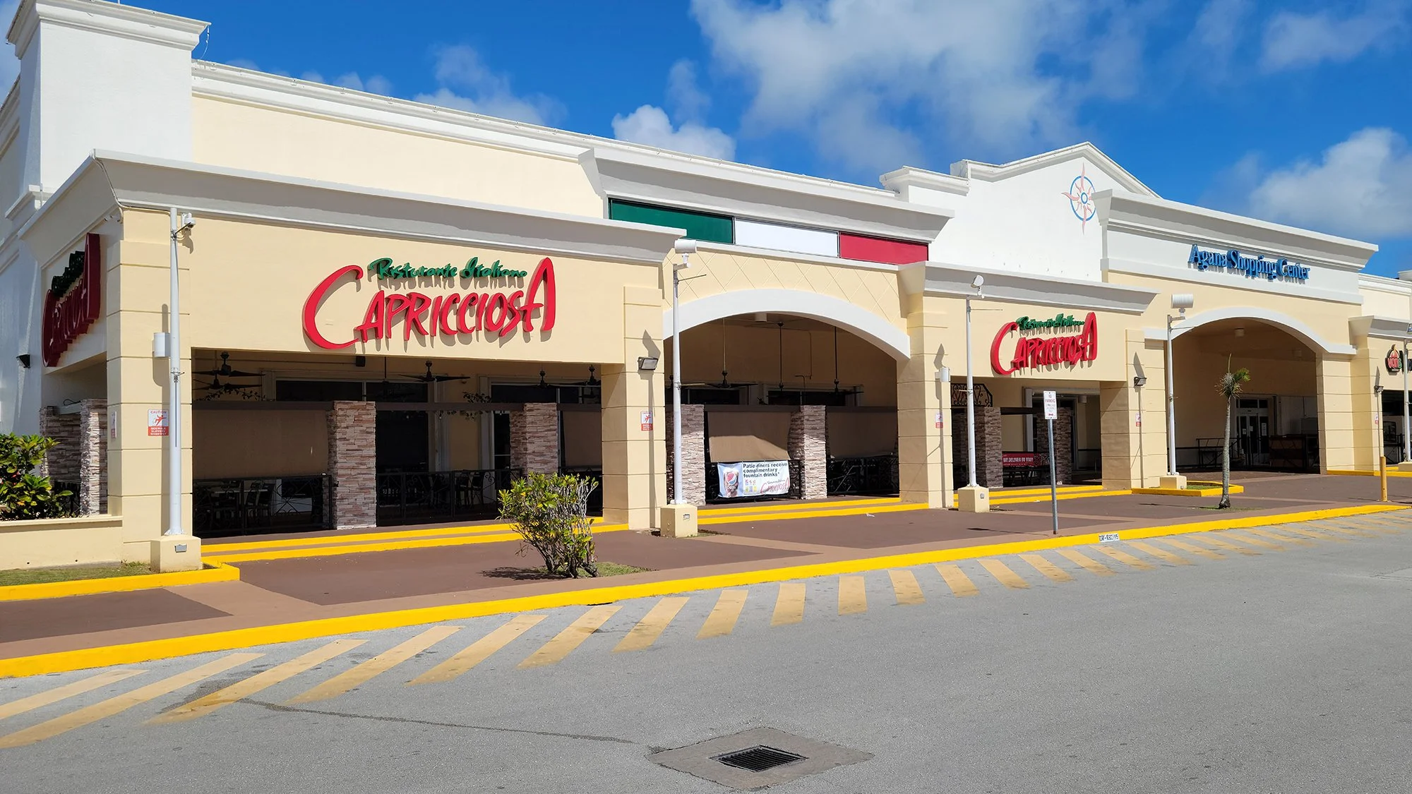 Shopping plaza with a restaurant called Capricciosa and an Argana shopping center entrance, with yellow parking lines and a small bush in the foreground.