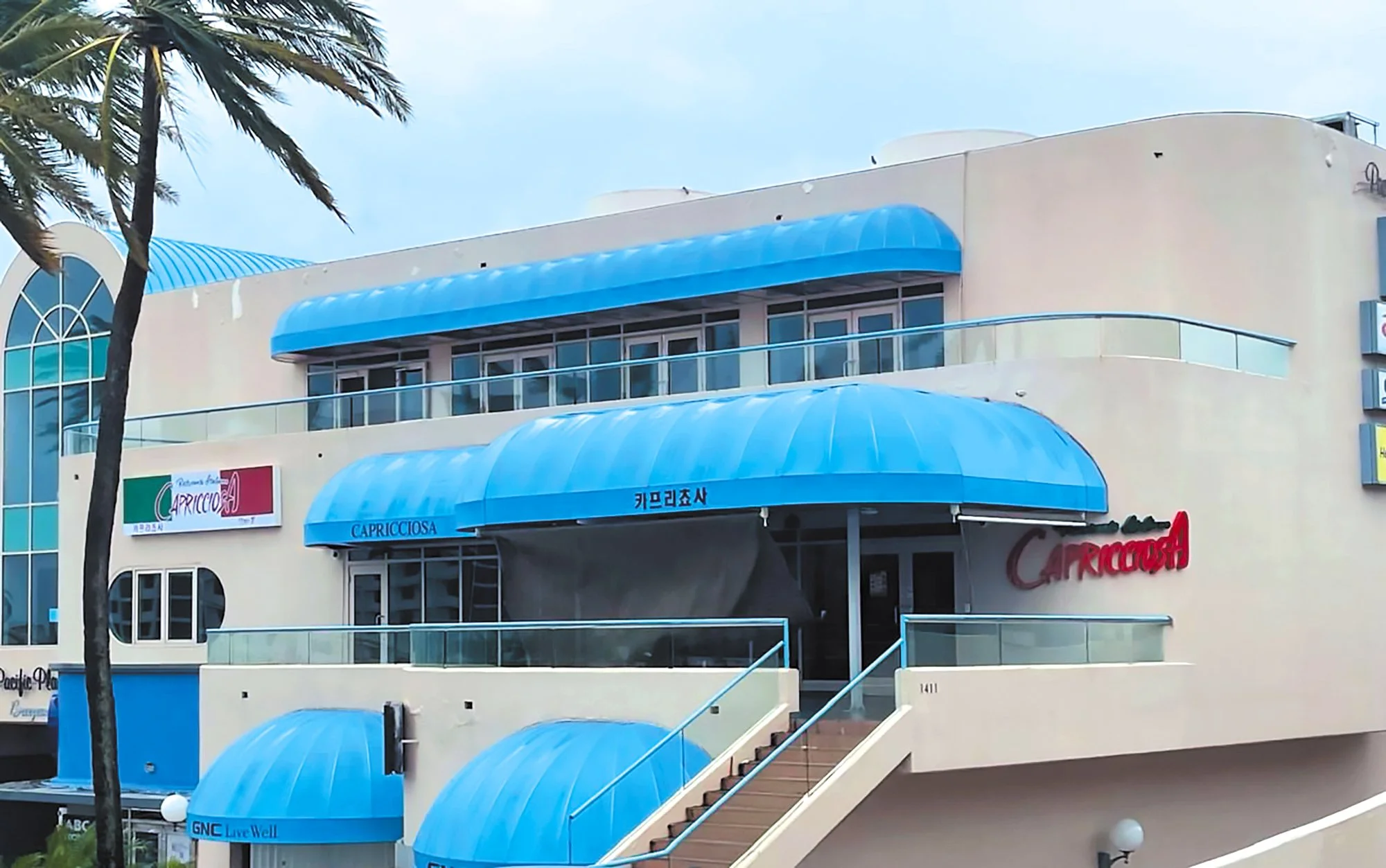 A modern, multi-story building with exterior blue awnings and curved glass railings. There are signs on the building with the name 'Capricciosa' and other storefront signs.