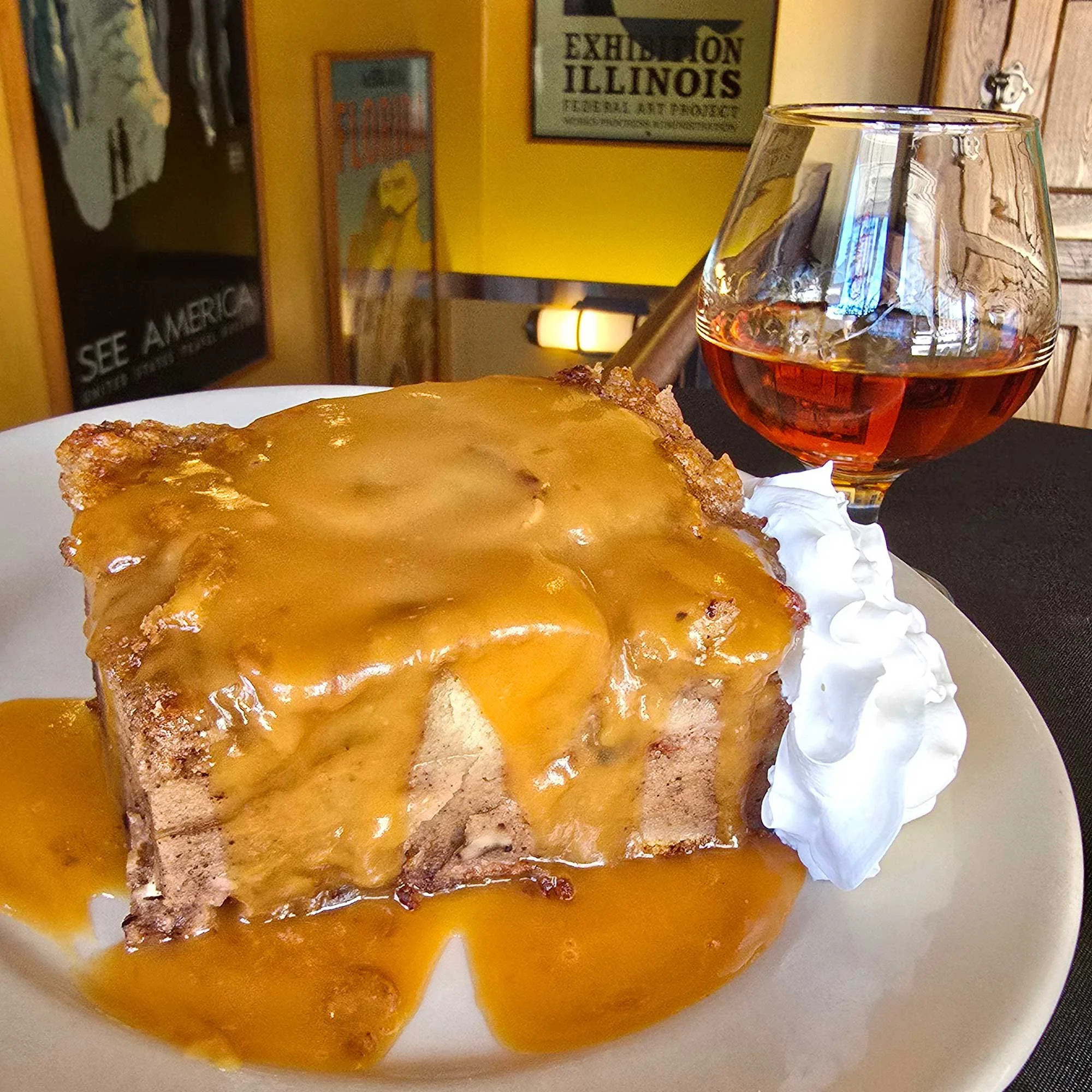 A slice of cherry pie with caramel sauce and whipped cream, served with a glass of rosé wine in a diner or cafe setting.
