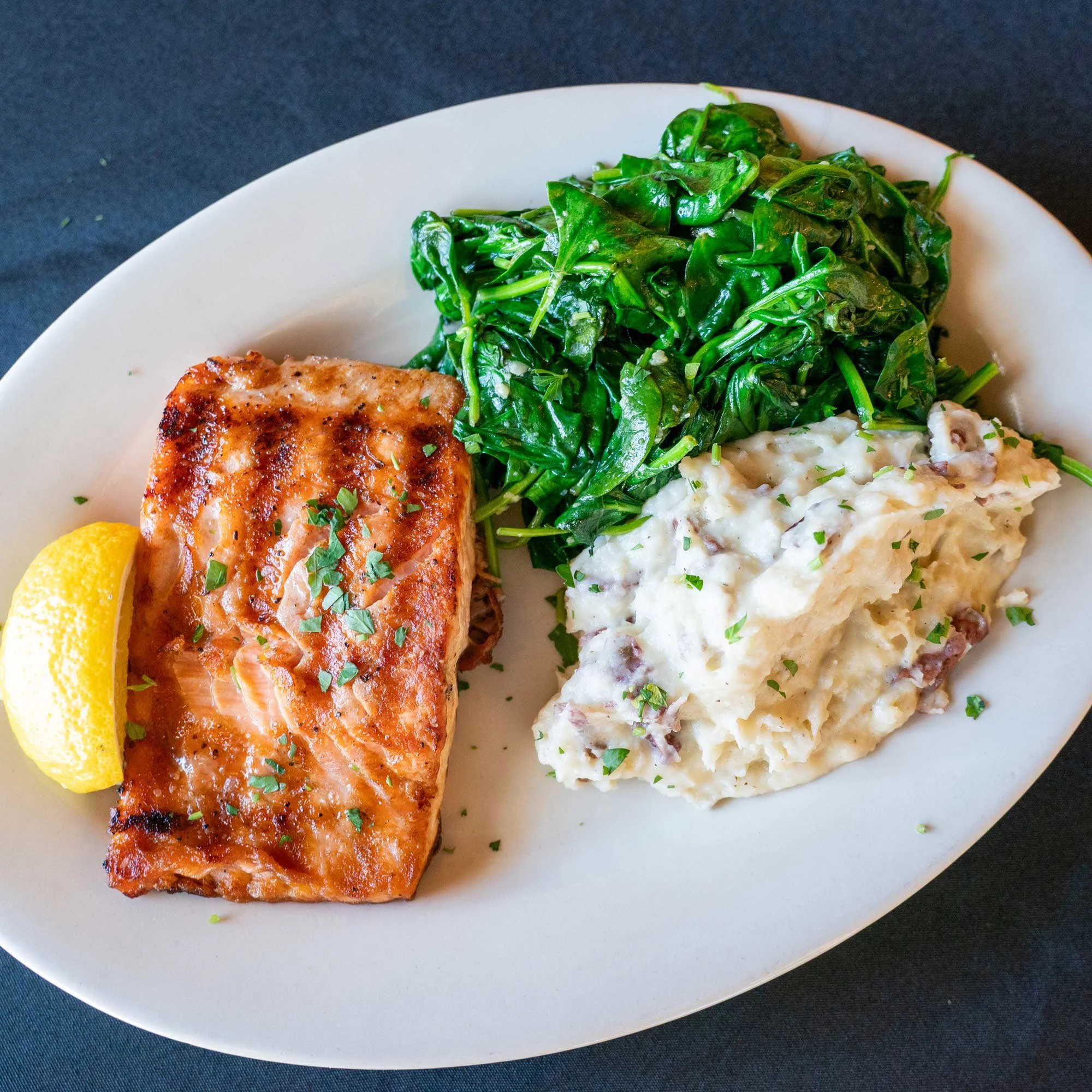 Plate with cooked salmon steak garnished with chopped herbs, lemon wedge, sautéed spinach, and mashed potatoes with herbs.