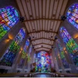 Interior view of a church or cathedral with large stained glass windows and wooden ceiling beams.