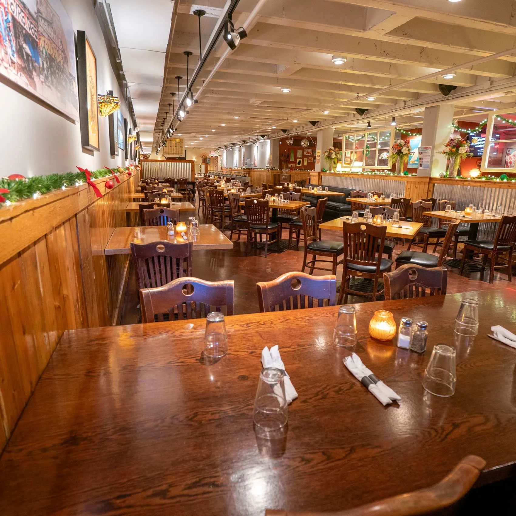 Empty restaurant with wooden tables and chairs decorated for the holidays, with candles on the tables and holiday garlands and flowers along the walls.