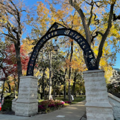 Entrance arch with metal sign in a park during fall, surrounded by colorful autumn trees.