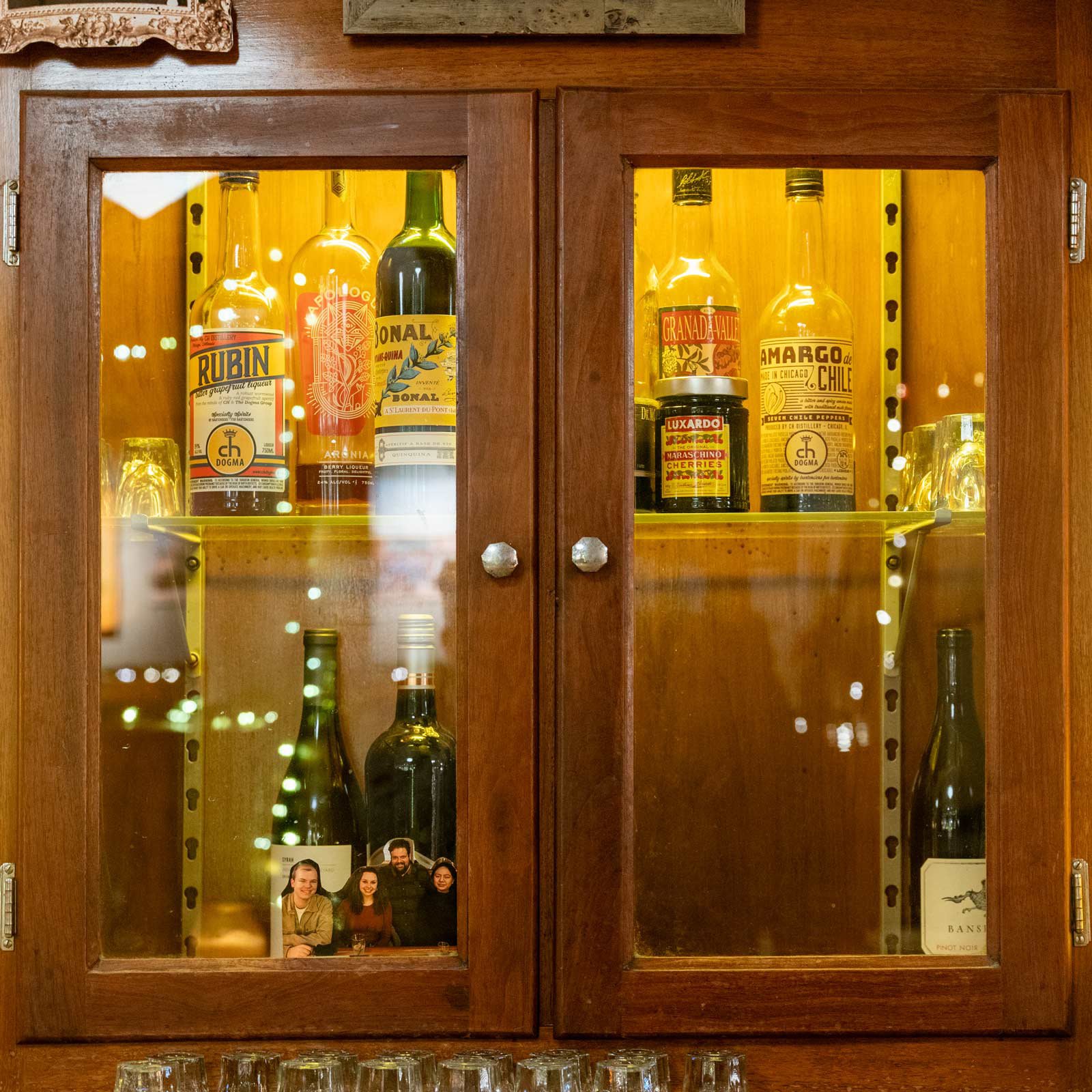 A wooden cabinet with glass doors containing bottles of alcohol, including wine and spirits, with a reflection of fairy lights and a small photo of four people inside the cabinet.