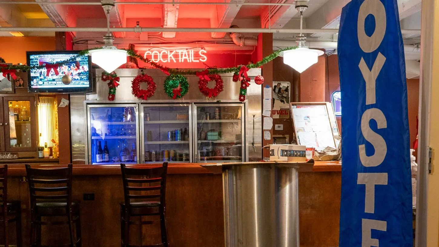 Bar area decorated with red and green Christmas garland and wreaths, with a neon sign reading 'Cocktails' above, and a large blue 'OUTSIDE' sign to the right.