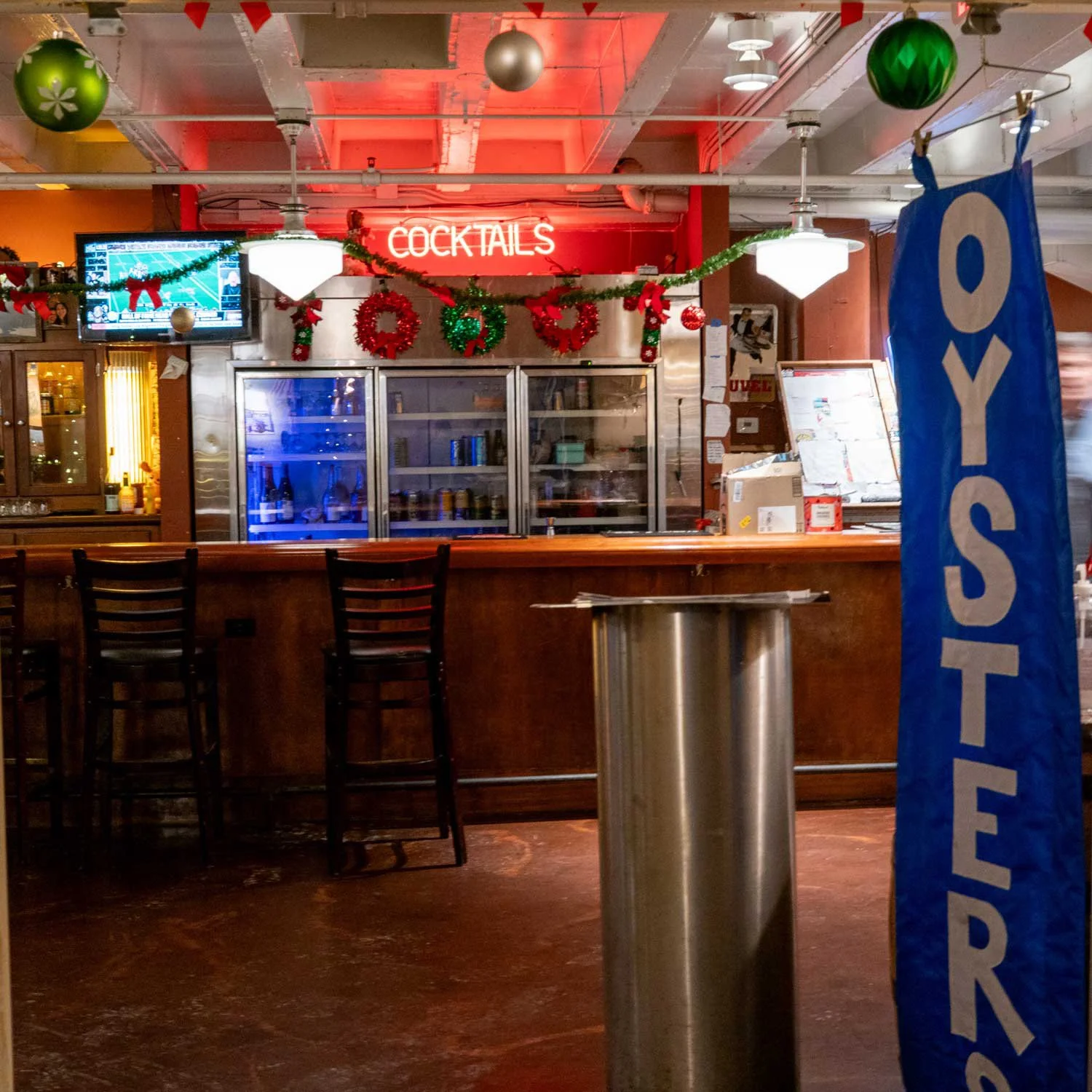 Bar counter decorated with Christmas ornaments, including green and gold baubles and holiday wreaths, with a neon sign reading 'COCKTAILS' and a blue 'YES! ORDER' stand in the foreground.