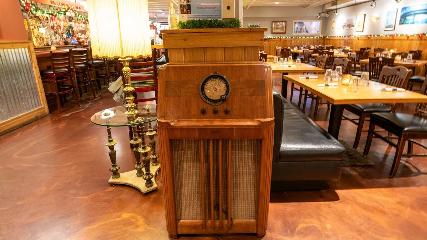 Vintage wooden radio standing in a restaurant or cafe with wooden tables and chairs, a glass side table with brass candle holders, and warm lighting.