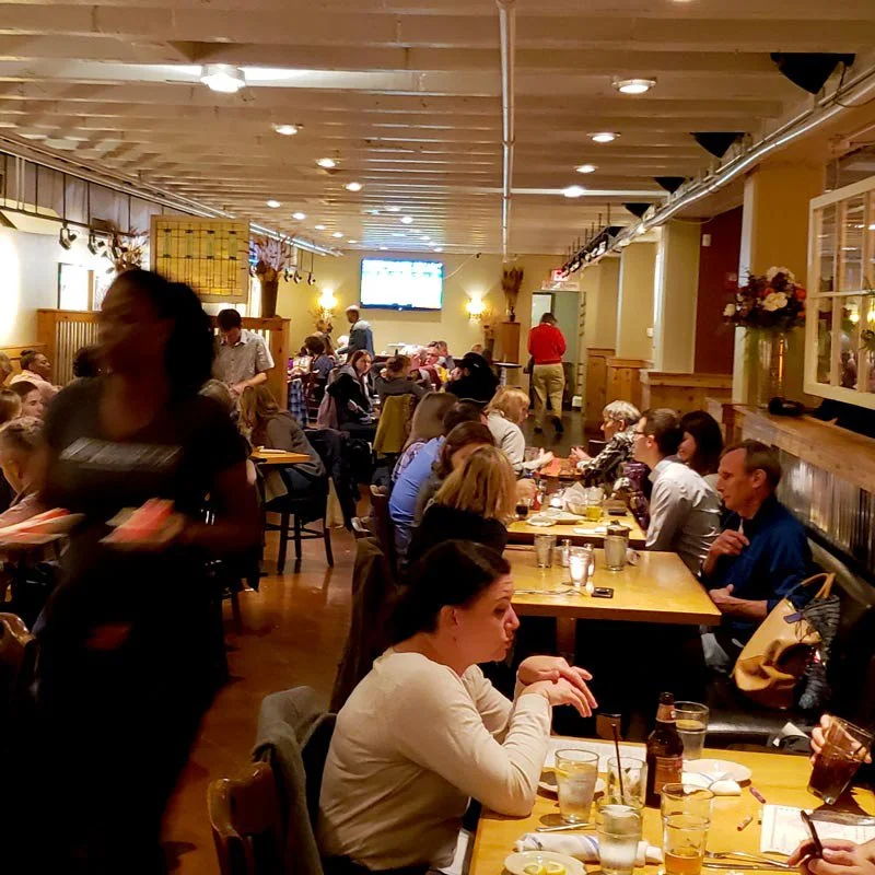 A busy restaurant with patrons dining at wooden tables, servers attending to guests, and a television mounted on the wall in the background.