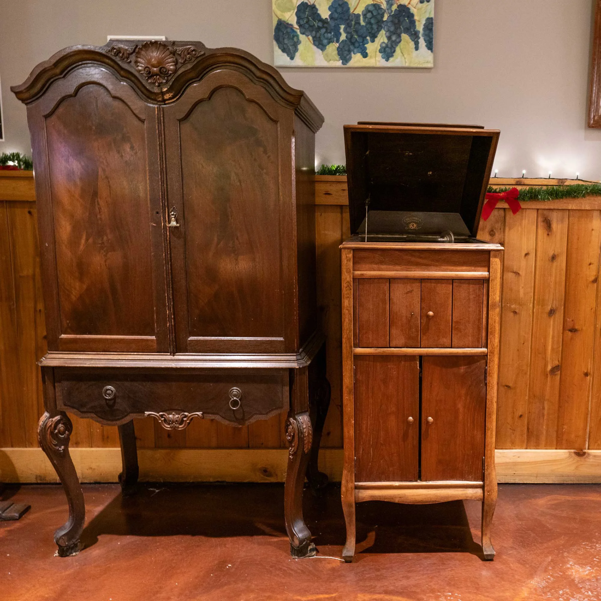 Two antique wooden furniture pieces, including a large armoire on the left with intricate carvings and curved legs, and a smaller wooden cabinet with a record player on top, against a wooden-paneled wall with a grape and leaf artwork above.
