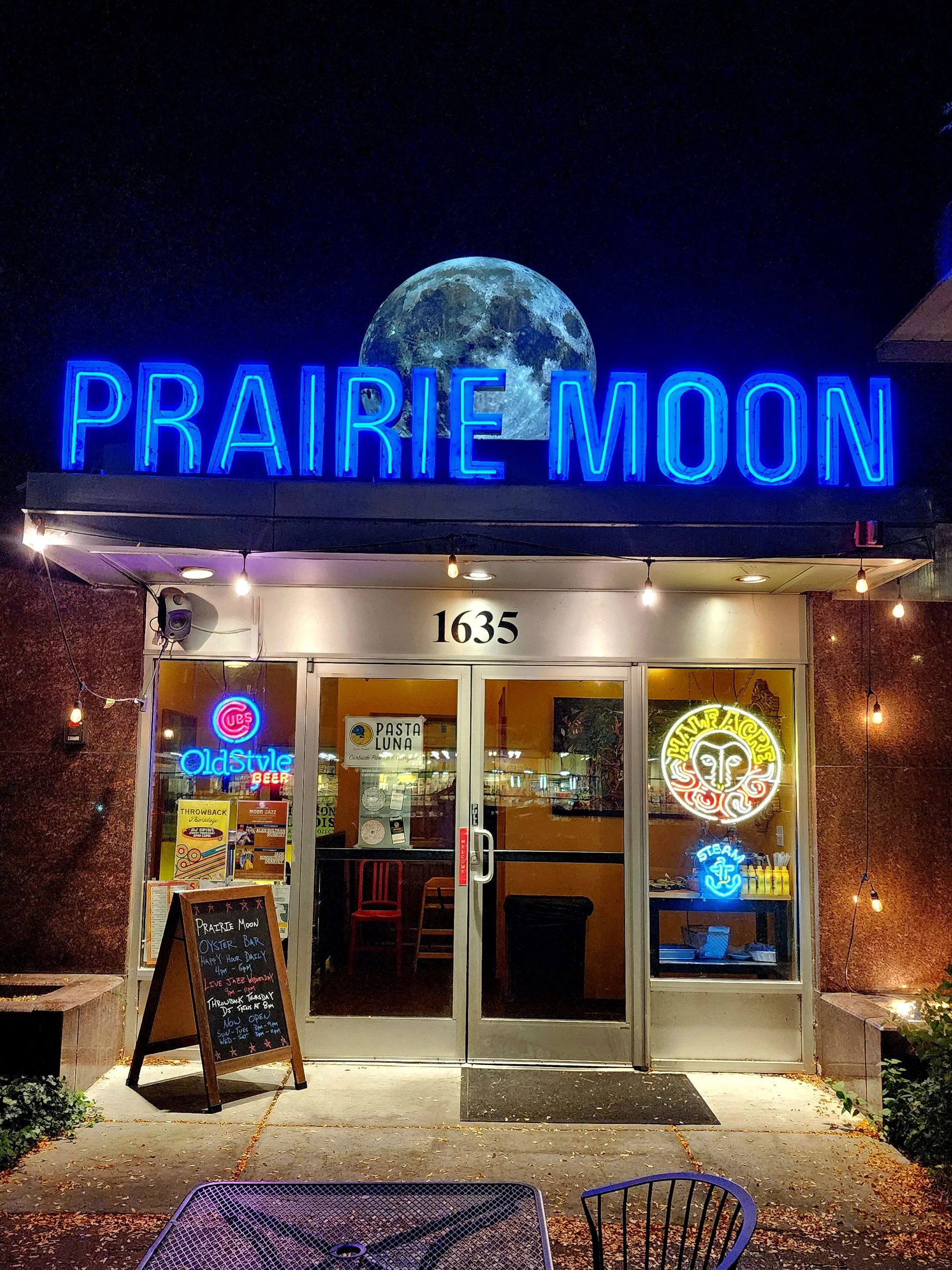 Night view of the Prairie Moon restaurant with a neon sign above the entrance, featuring a moon and the restaurant's name. Additional neon signs display 'Old Style' beer, 'Hawaiian' style, and 'Steam'. The entrance has a blackboard menu outside, and 