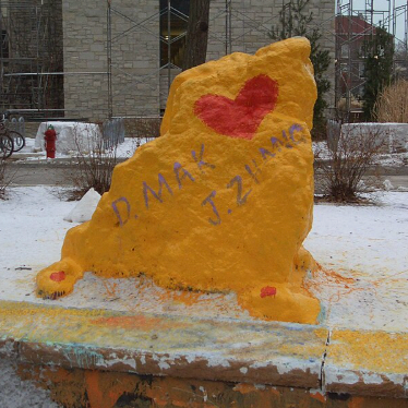 Yellow sculpture of a dog with red heart painted on its side and handwritten names 'D. Mack' and 'J. Ziam' in purple, outdoors on a snowy day.