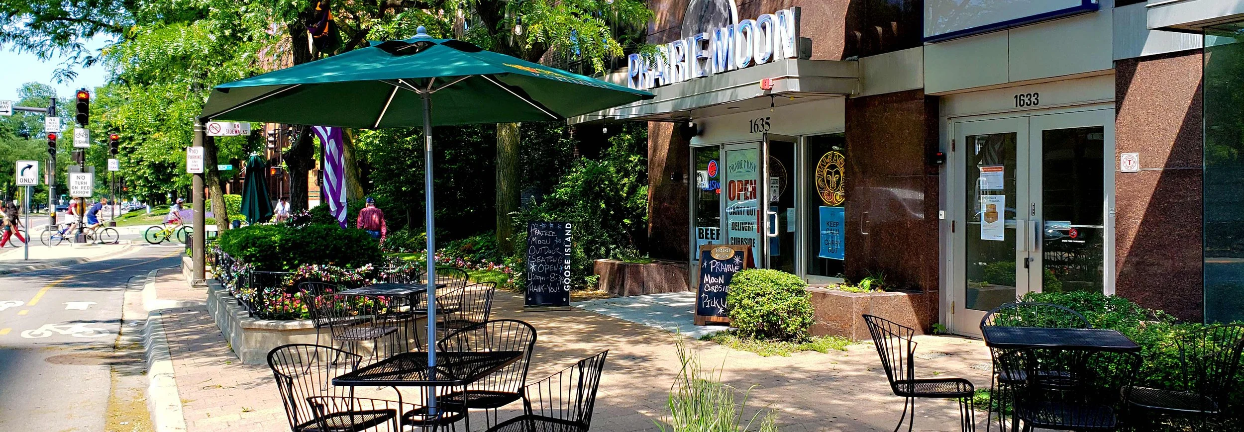 Street view of Prairie Moon restaurant outdoor seating, black chairs, and tables under a large green umbrella. The restaurant has signs indicating it is open, and people are seen walking and biking in the background. Trees and greenery surround the a