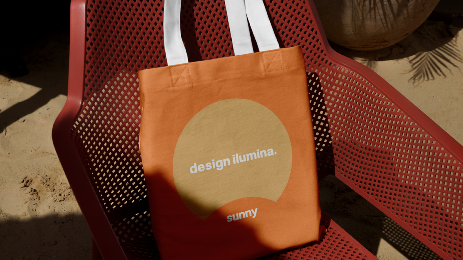 Sacola de pano laranja com inscrição "design ilumina. sunny" sobre uma cadeira vermelha com suporte de malha em um ambiente ao ar livre.