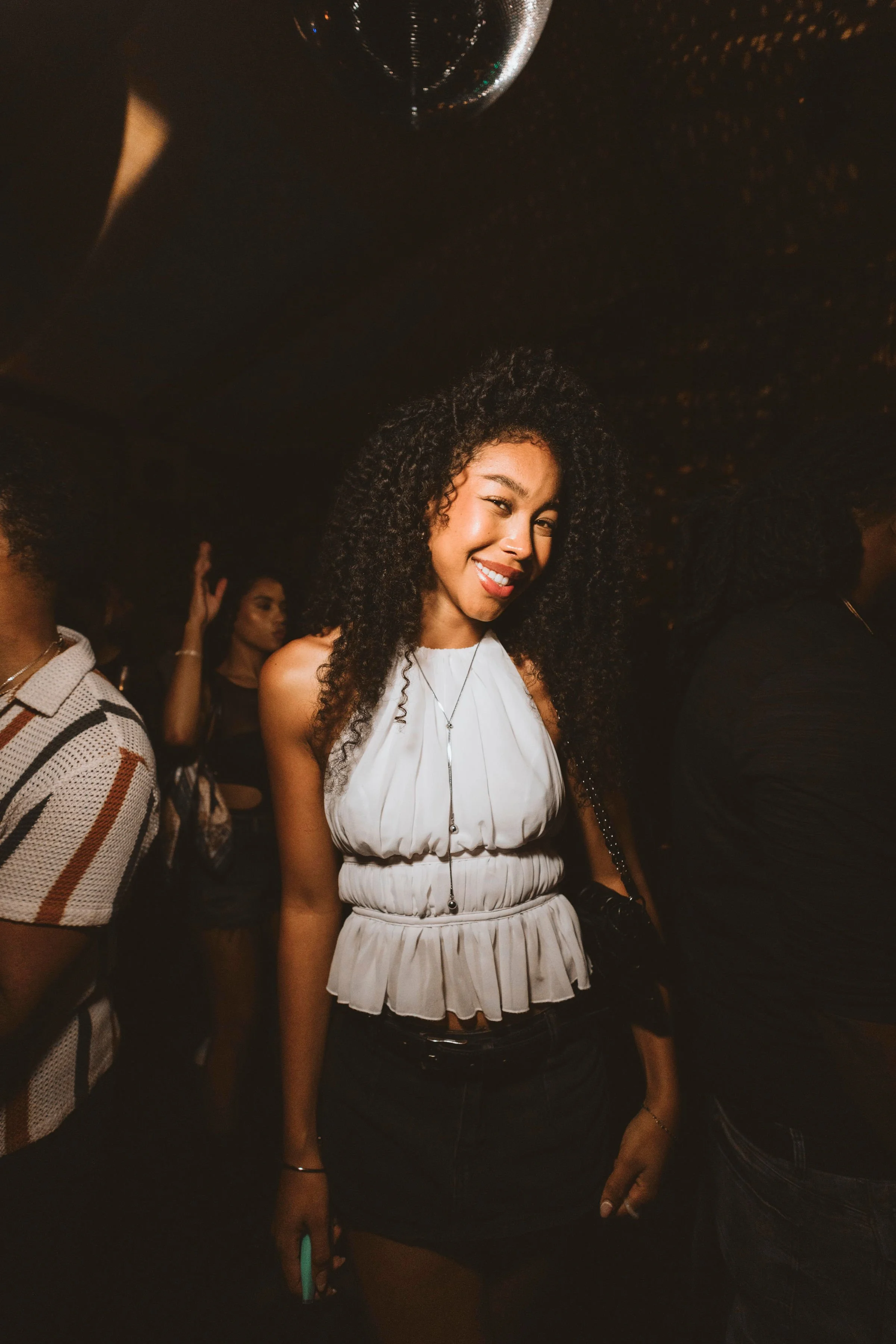 A young woman with curly hair smiling and winking at the Casa de Afro party by Dr. ADO in Mexico City, wearing a white sleeveless top and black skirt, surrounded by other people.