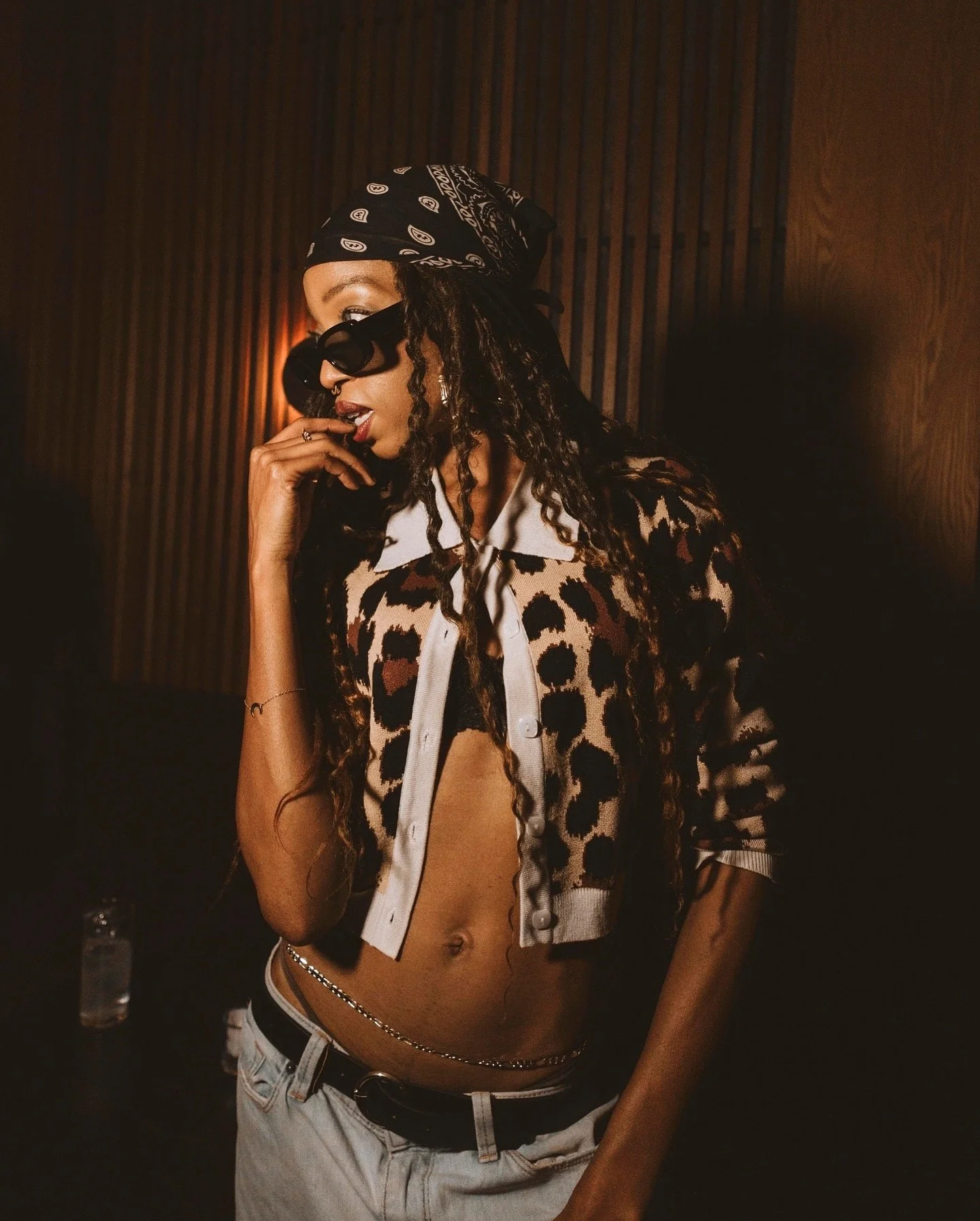 A woman with long, curly hair wearing sunglasses, a black bandana, a leopard print shirt, drawstring jeans, and a belly chain, posing against a dark wooden wall with warm lighting at a Casa de Afro party by Dr. ADO in Mexico City.