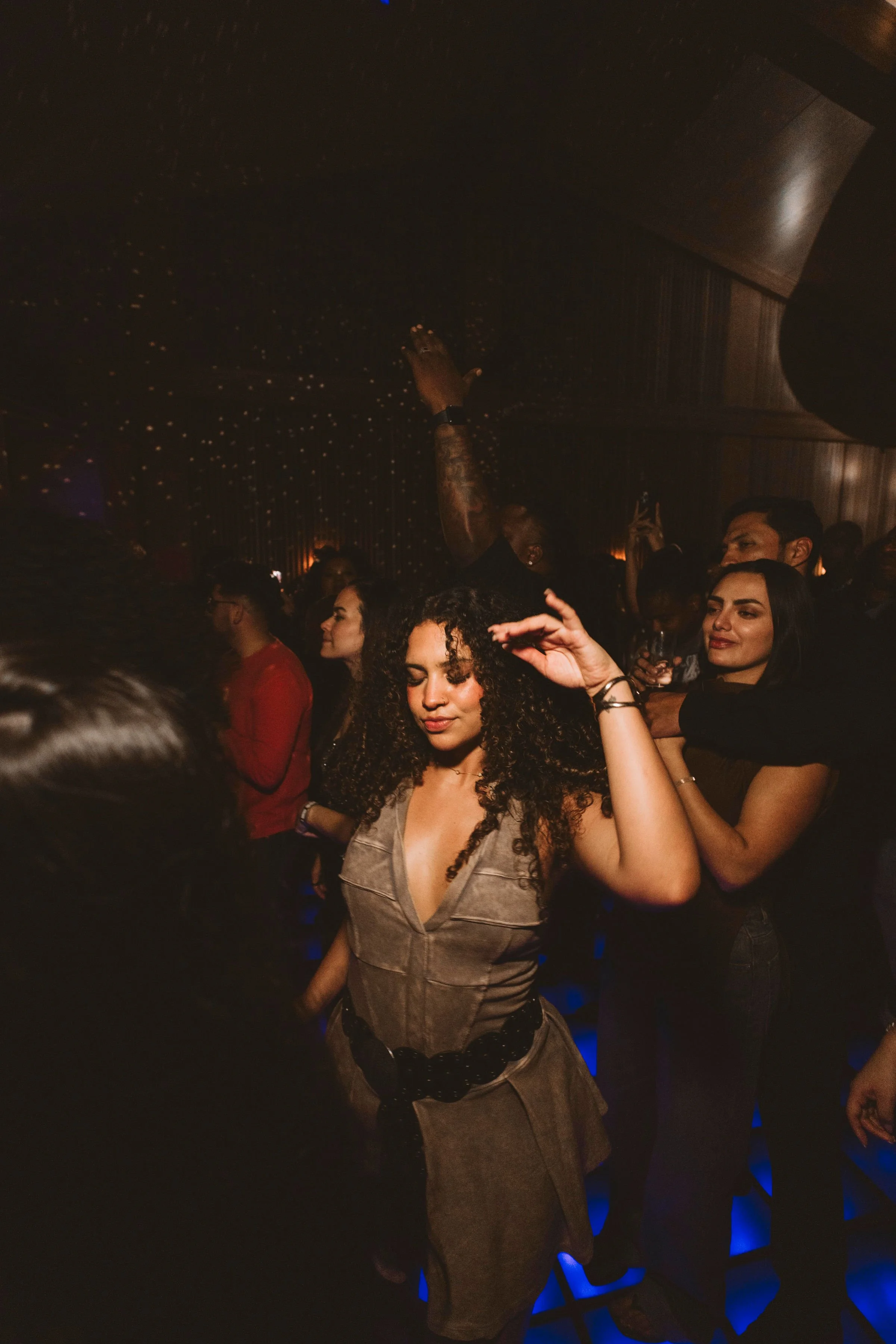 A woman with curly hair dancing with eyes closed in a dark, crowded nightclub with blue lighting. Dancing at a Casa de Afro party by Dr. ADO in Mexico City.