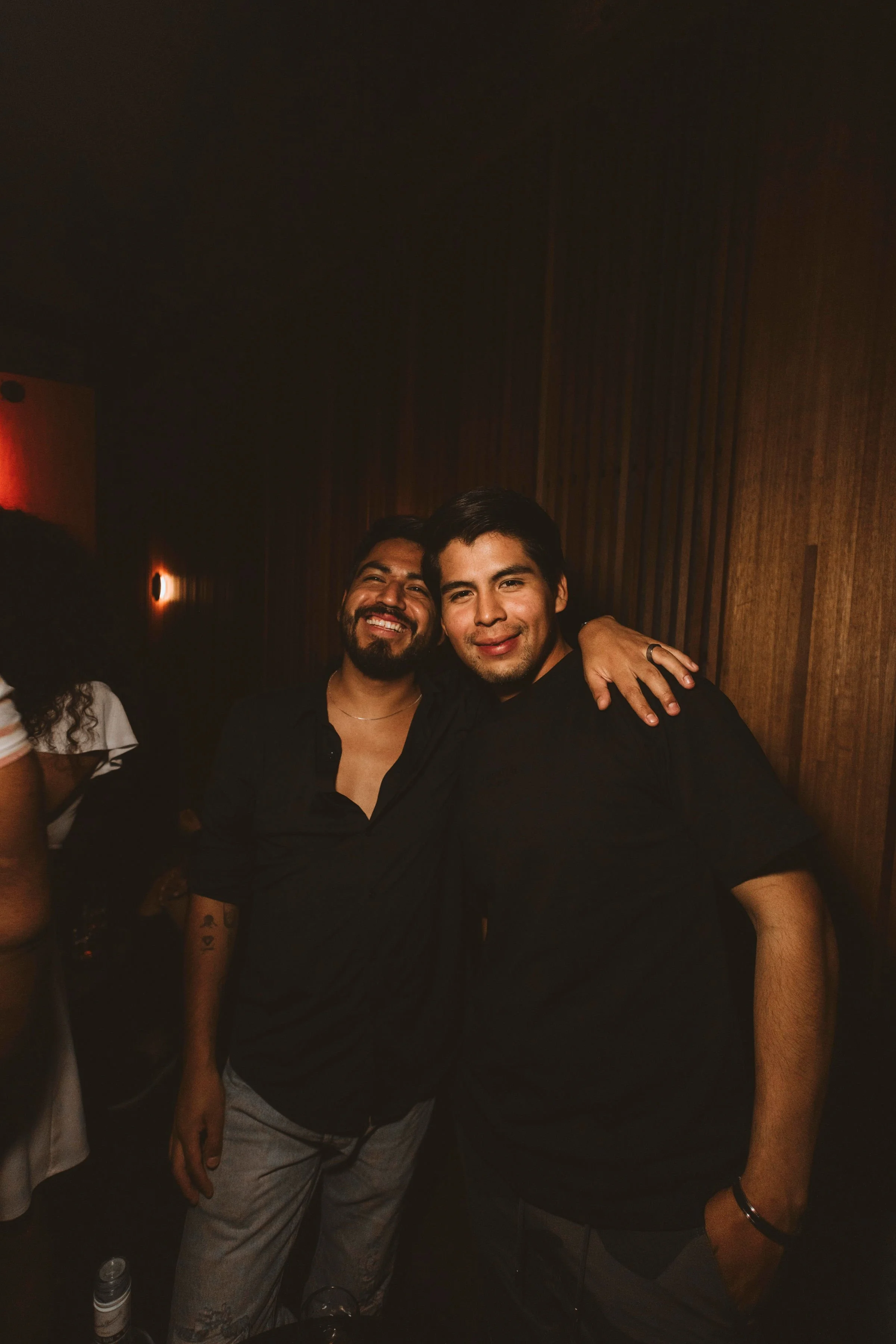 Two young men smiling and posing together indoors with a dark wooden wall background. One has a beard and is wearing a black shirt, the other has short dark hair and is also wearing a black shirt. The man on the left has his arm around the other's sh