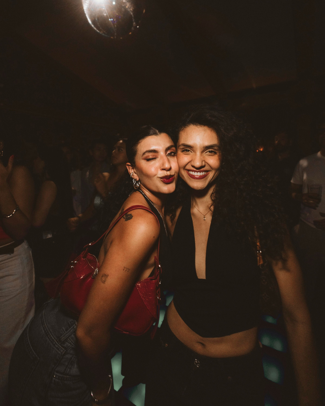 Two women smiling and posing together at a party or club, with a disco ball overhead and dim lighting in the background at a Casa de Afro party by Dr. ADO in Mexico City.
