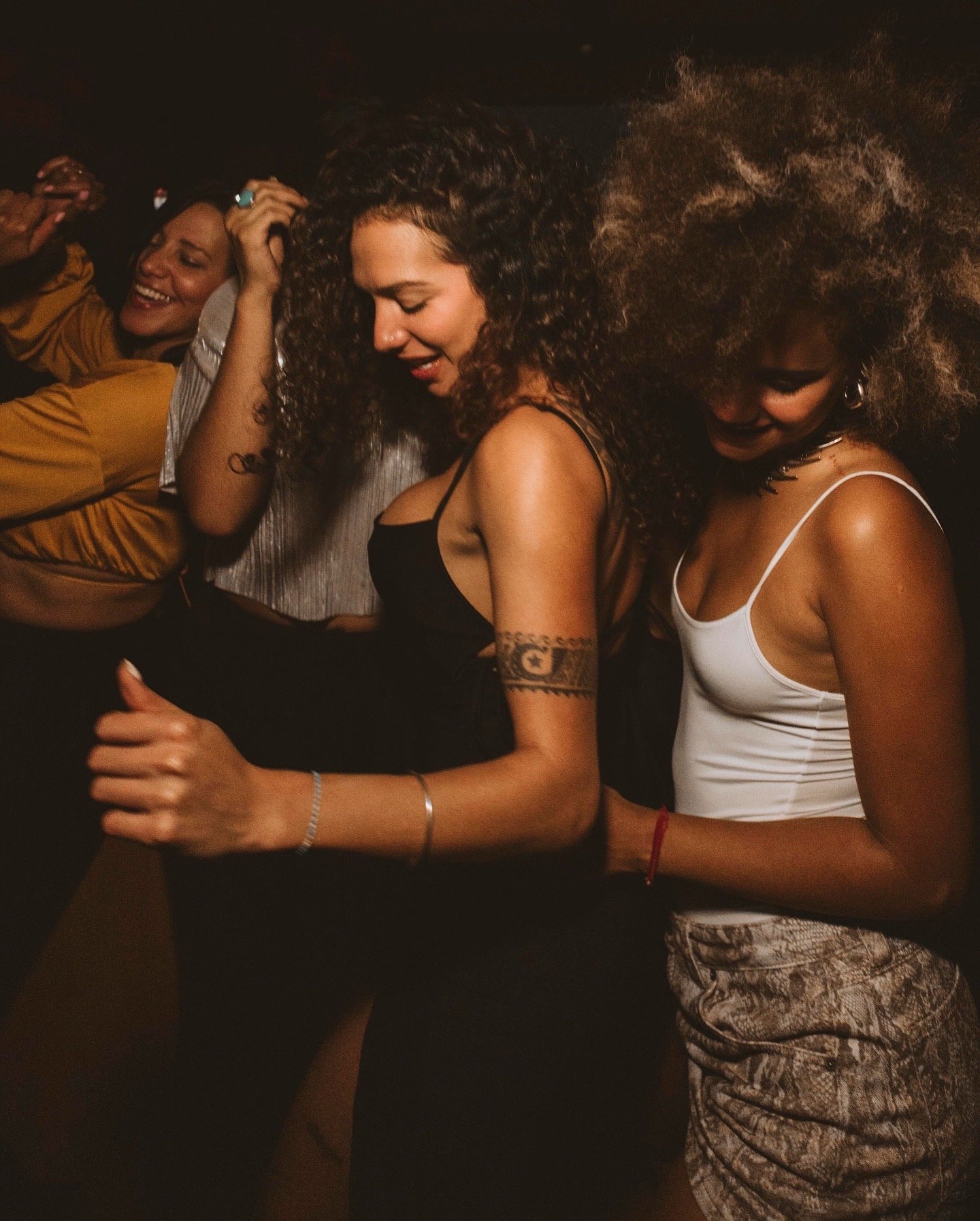 Three women dancing and smiling at a party, with dark background lighting at a Casa de Afro party by Dr. ADO in Mexico City.