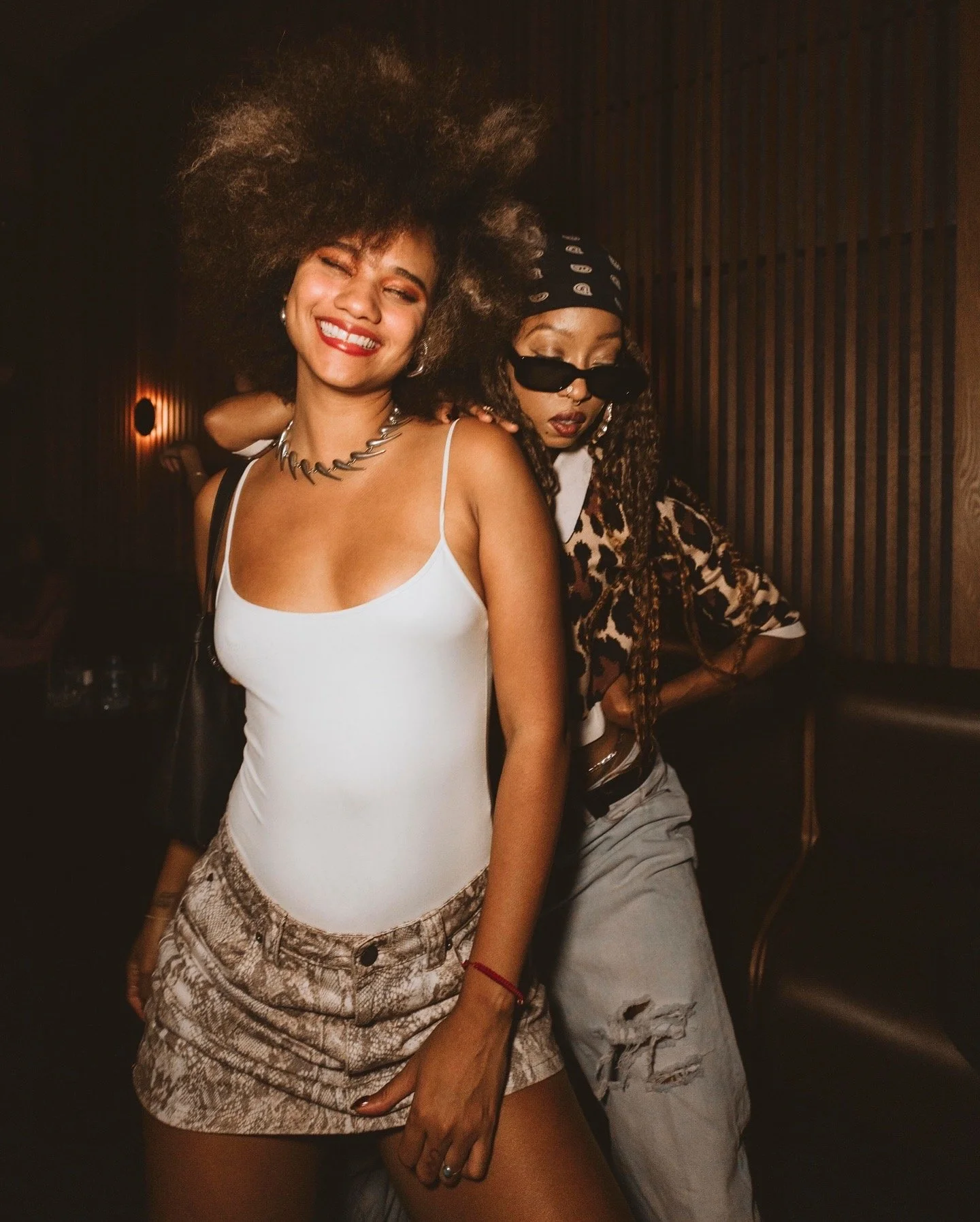 Two women dancing together at a Casa de Afro party by Dr. ADO in Mexico City, with one smiling brightly and the other wearing sunglasses and a bandana.