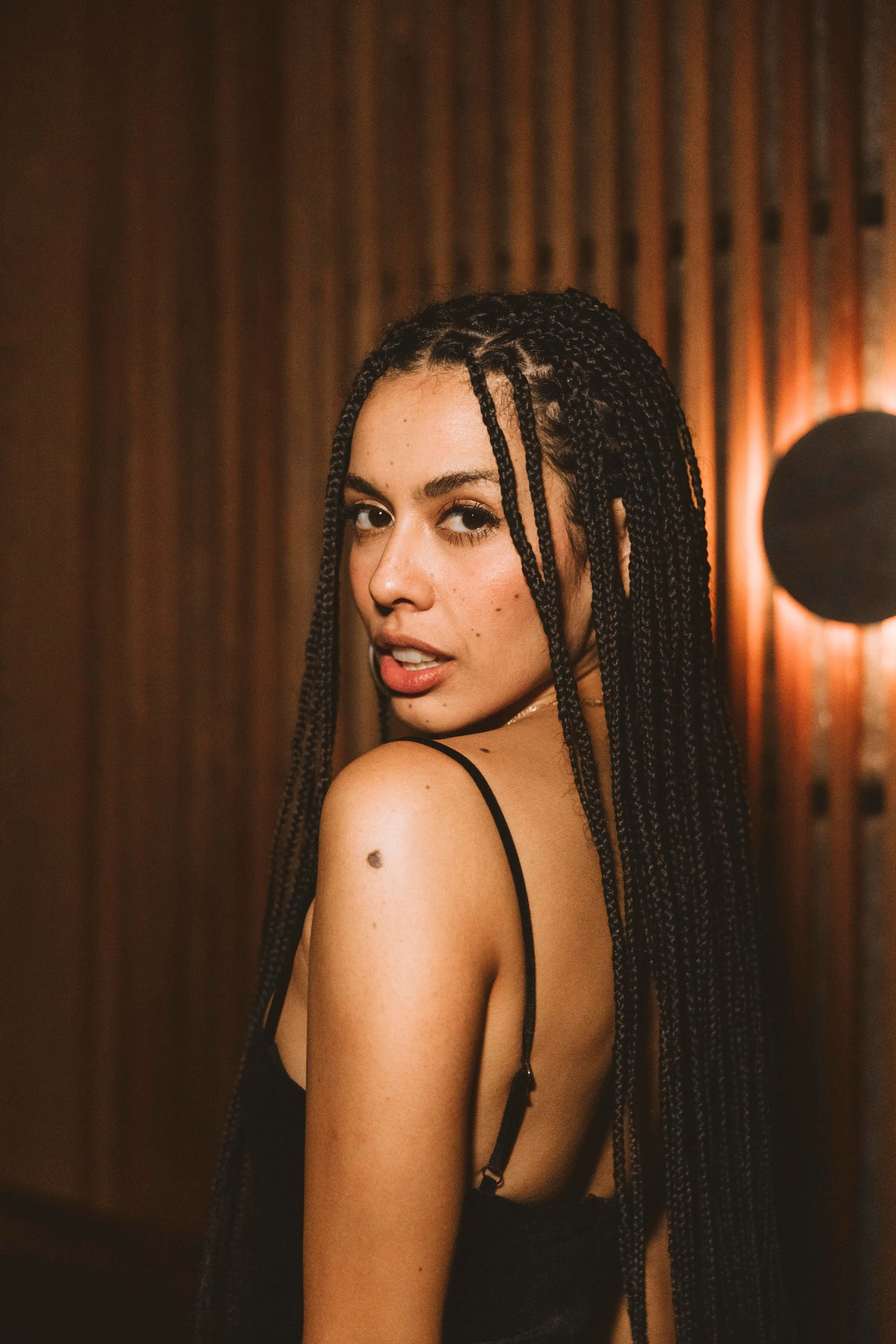 A young woman with long braided hair, wearing a black sleeveless top, posing against a wooden wall with a circular light fixture in the background at Casa de Afro by Dr. ADO in Mexico City.