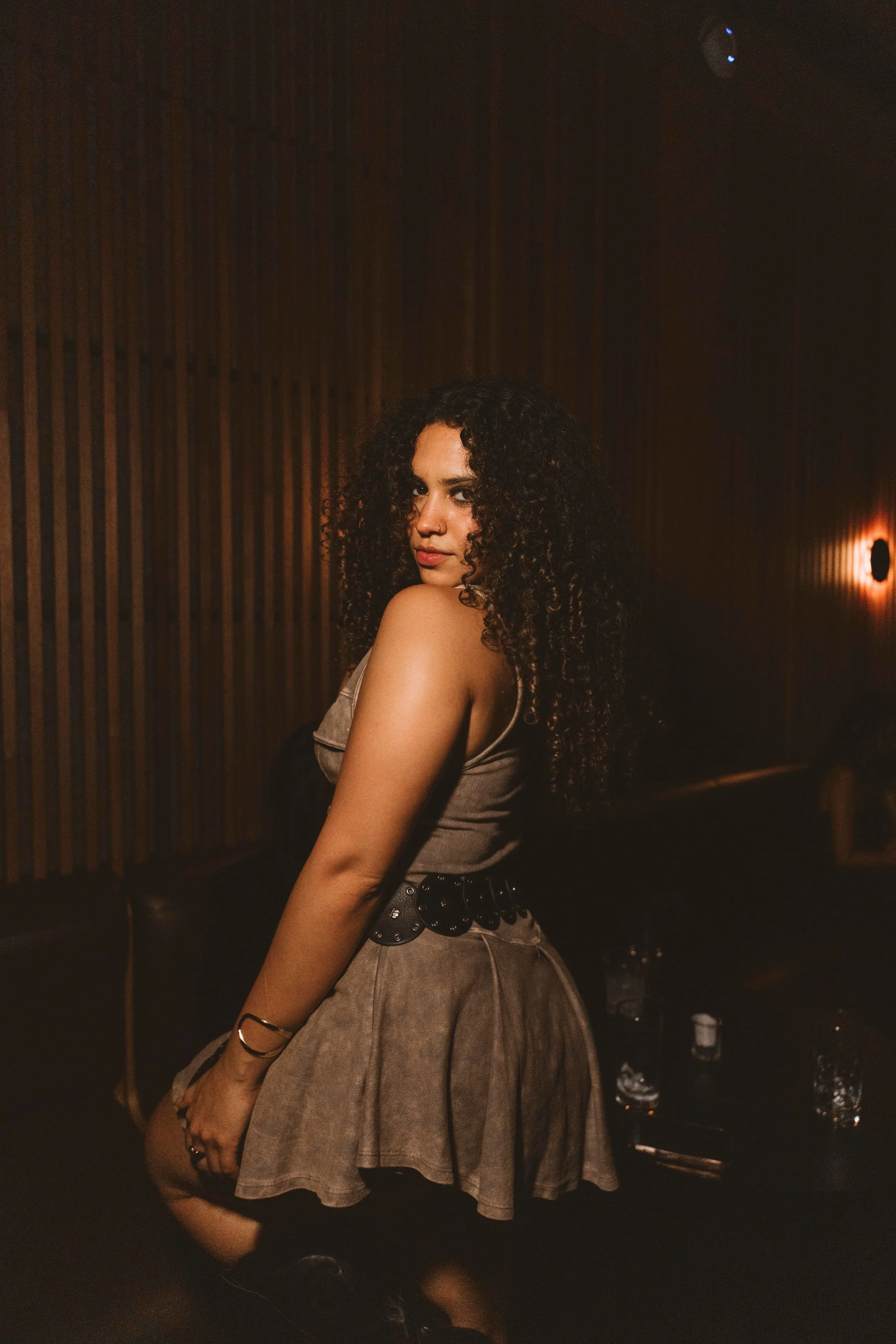 A woman with dark curly hair posing in a dimly lit room with wooden wall accents, wearing a beige sleeveless top, a black belt, and a matching skirt. Partying at Casa de Afro by Dr. ADO in Mexico City. 