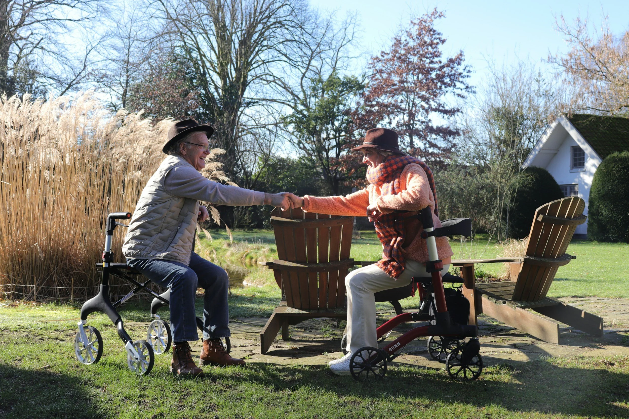 Two elderly women, both using mobility aids, shake hands and smile in a sunny garden with trees, a white house, and wooden outdoor furniture.