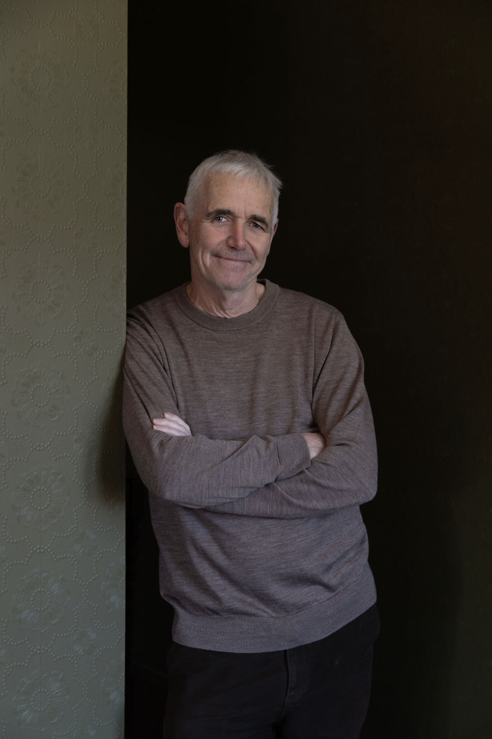 A man with gray hair and a slight smile, wearing a brown sweater, standing with arms crossed against a dark background.
