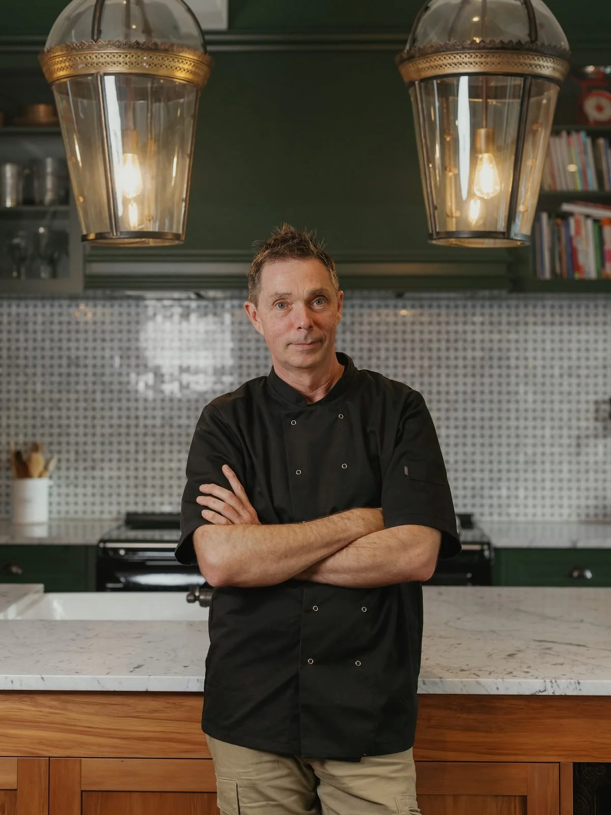A male chef stands with arms crossed in a modern kitchen with a white marble countertop, wooden cabinets, and backsplash tiles. Large pendant lights hang from the ceiling above.