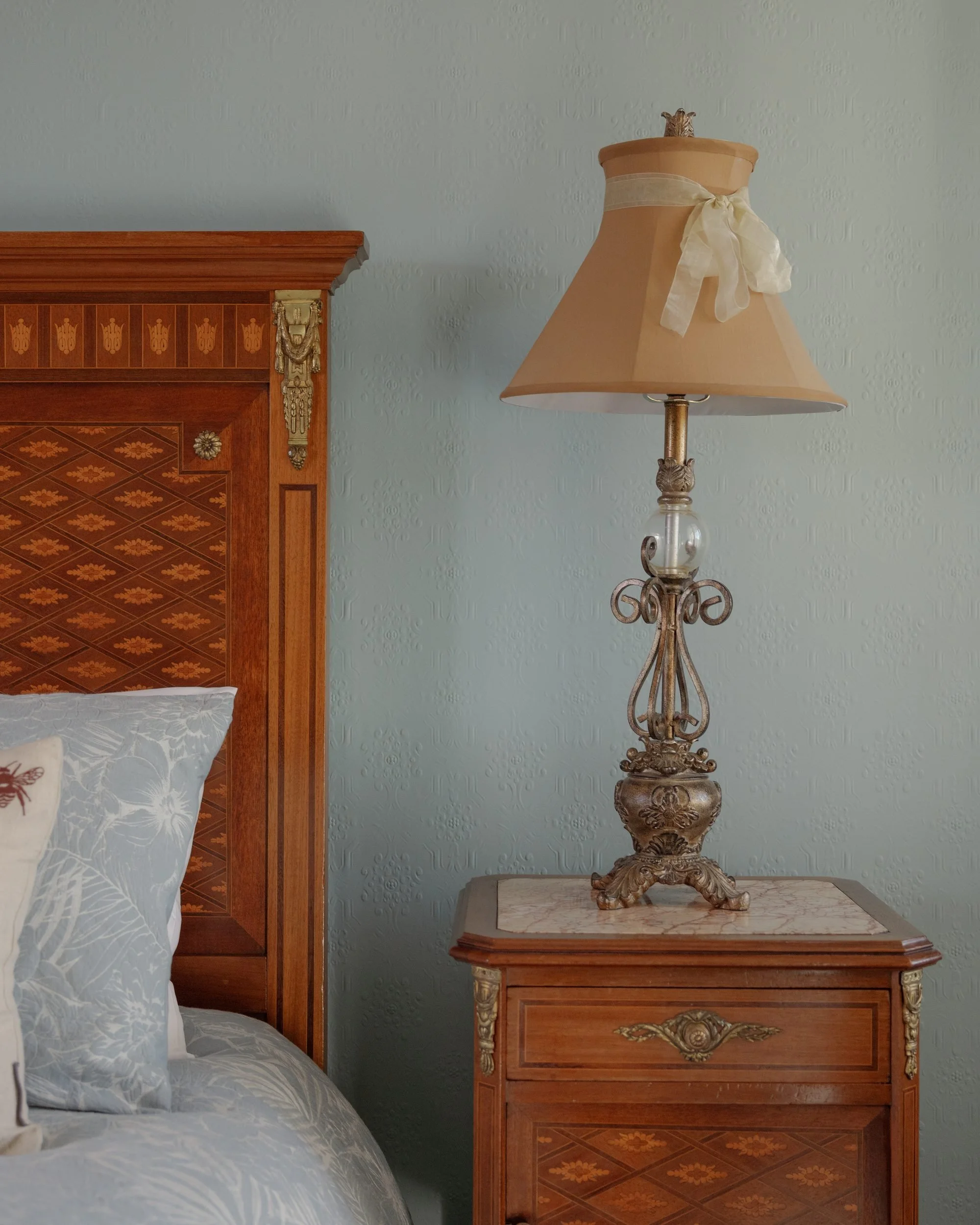 A bedroom scene with a large wooden headboard, a decorative side table with an ornate lamp, and a bed with white and patterned pillows.