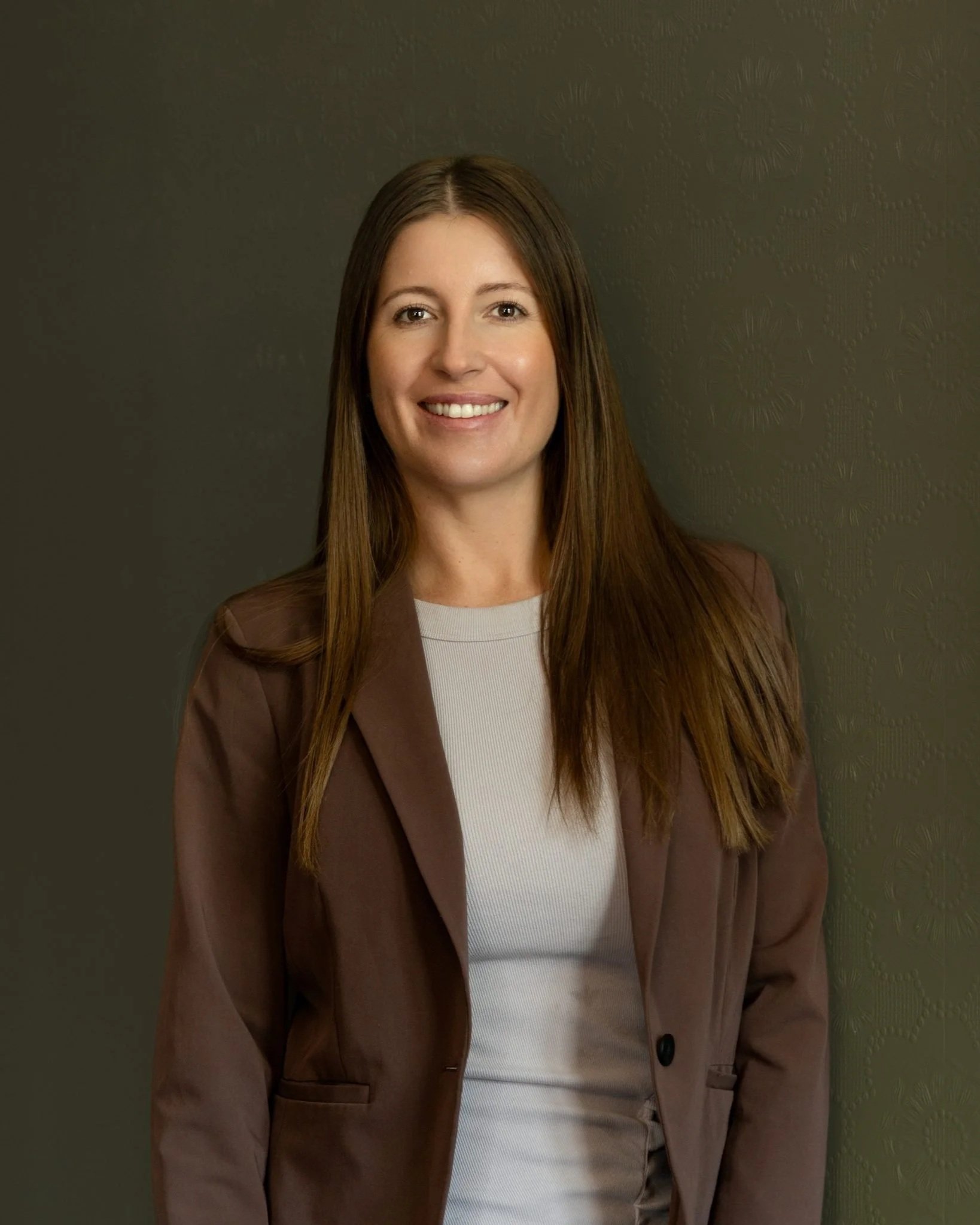 A woman with long brown hair, smiling, wearing a brown blazer over a white top, standing against a dark textured wall.