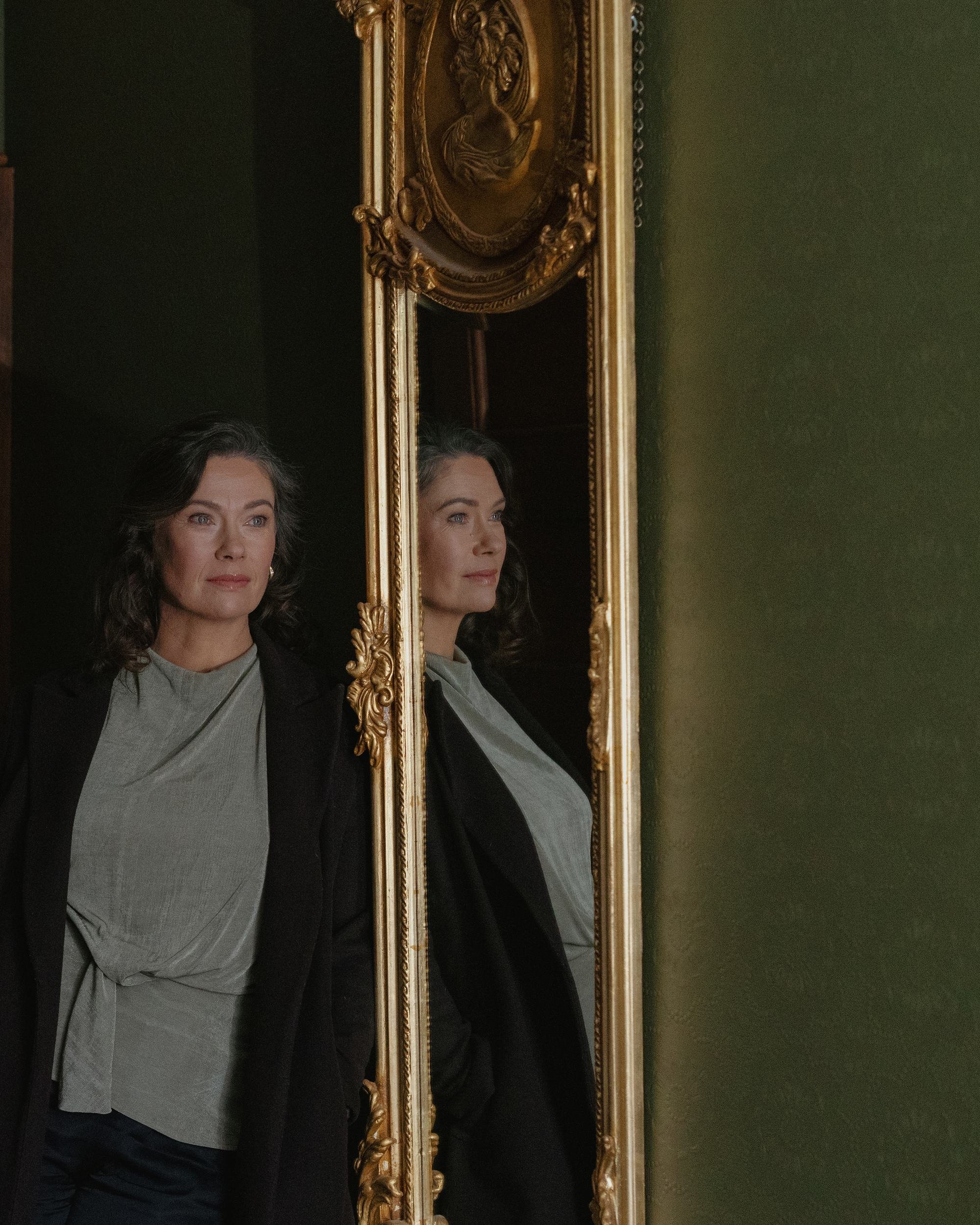 A woman with dark, wavy hair and light skin, wearing a black blazer and gray blouse, is looking at her reflection in a tall, ornate gold-framed mirror. The mirror is mounted on a dark wall, and there is a green wall to the right.