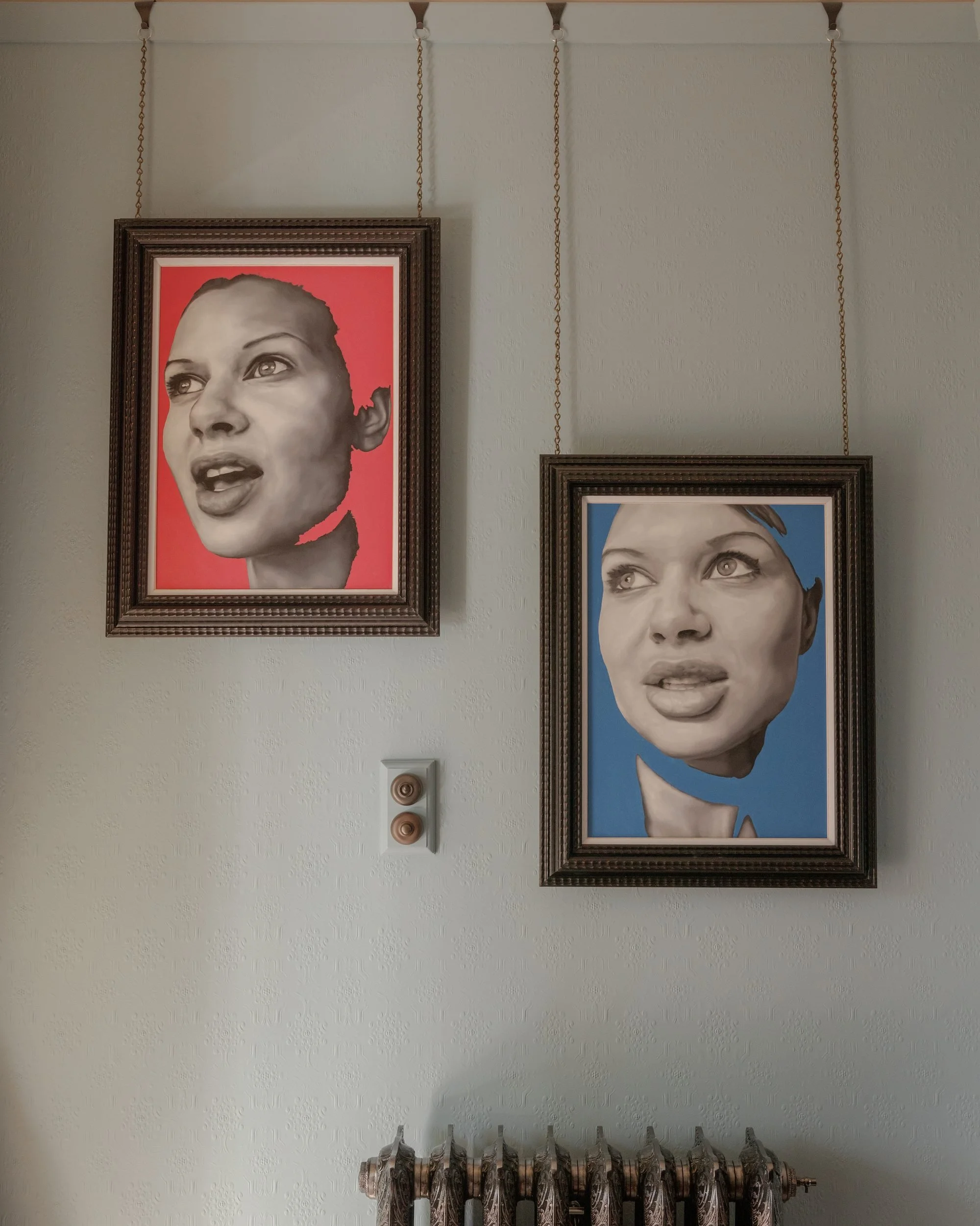 Two framed portrait paintings hanging on a light-colored wall, each showing a woman's face with different expressions. The left portrait has a pink background and the right with a blue background. Both are suspended by chains from hooks on the wall above.