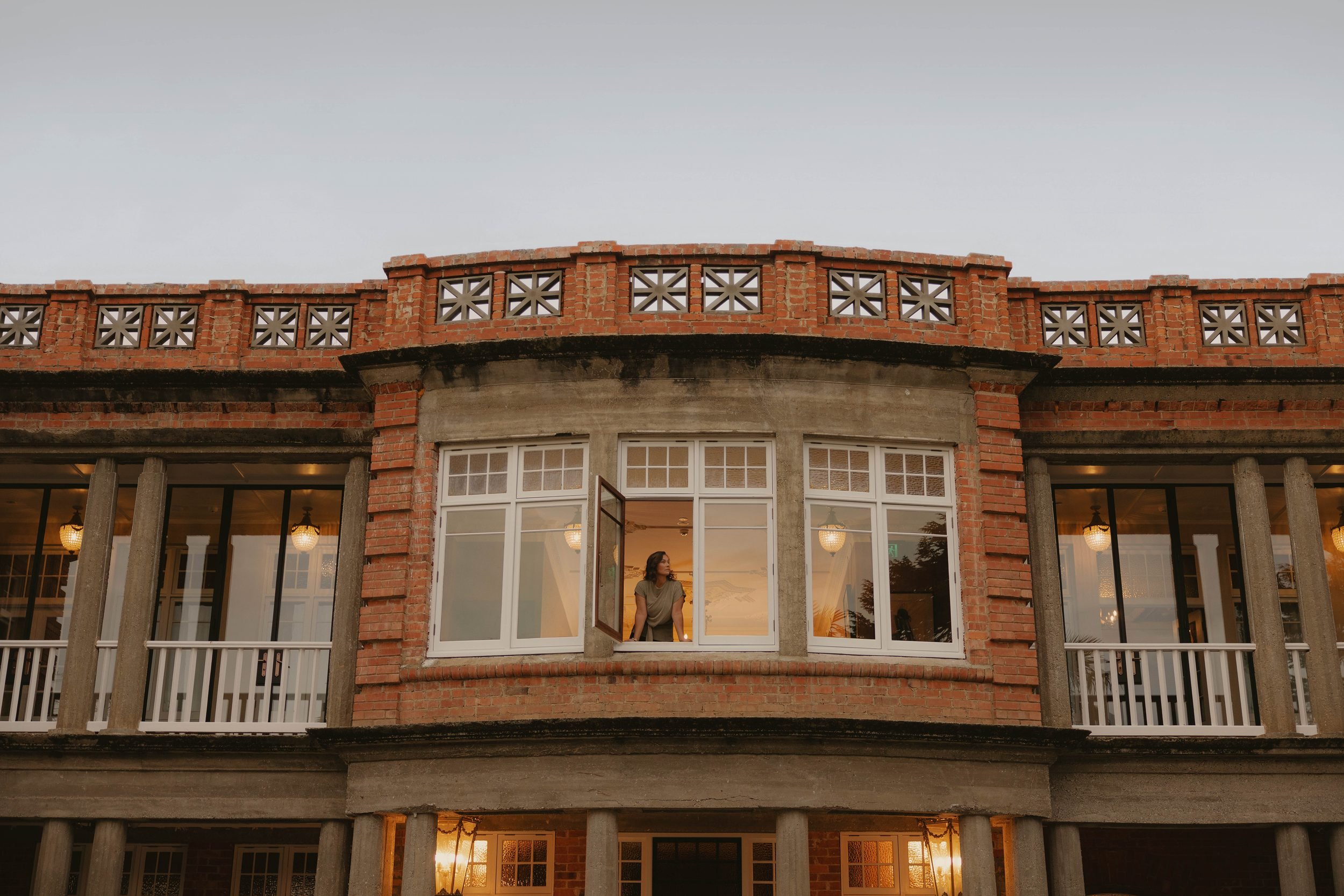 A woman standing inside a large window of a brick building, looking outside during sunset, with visible indoors lights and furnishings.